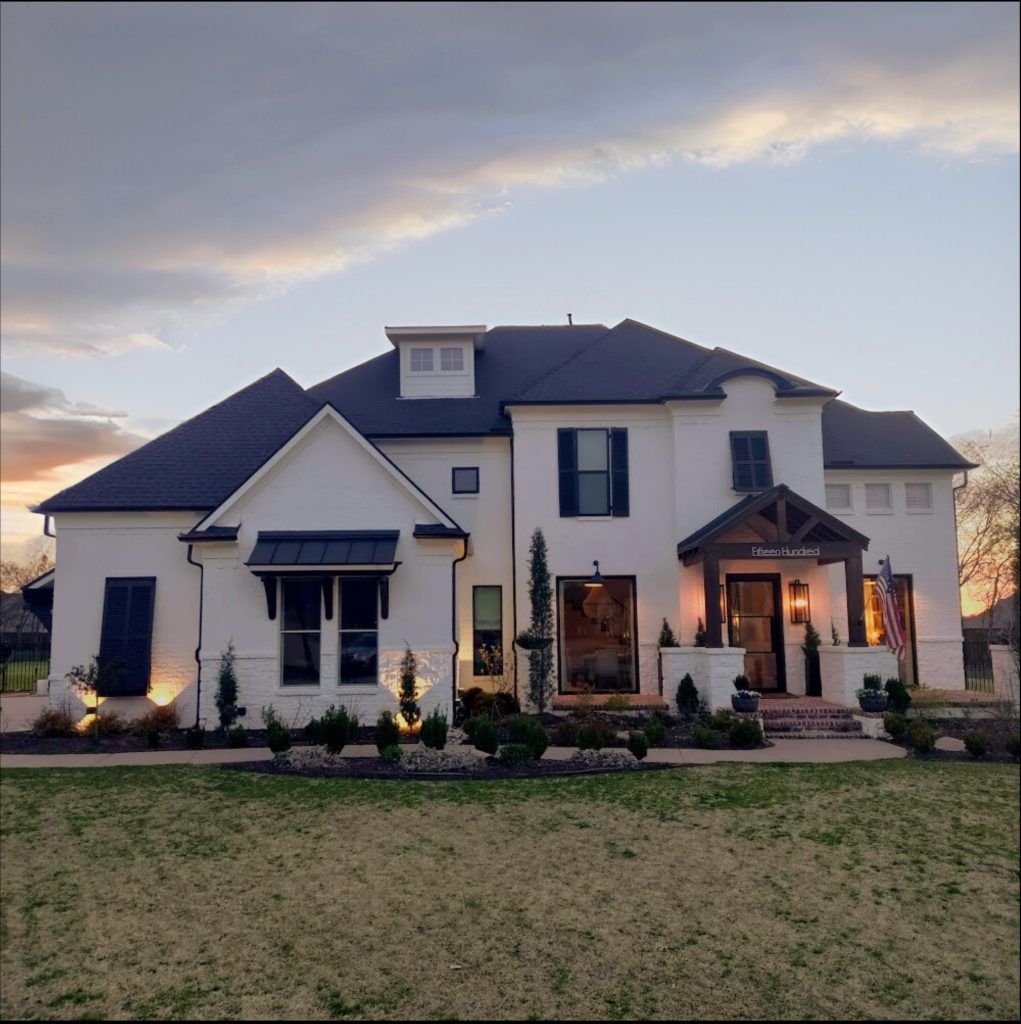 White two-story house with black shutters and door, brick porch, and landscaped yard at sunset.