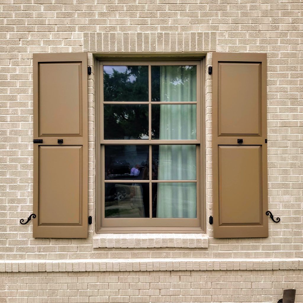 Window with open brown shutters on a brick wall. The window reflects a glimpse of a room and trees.