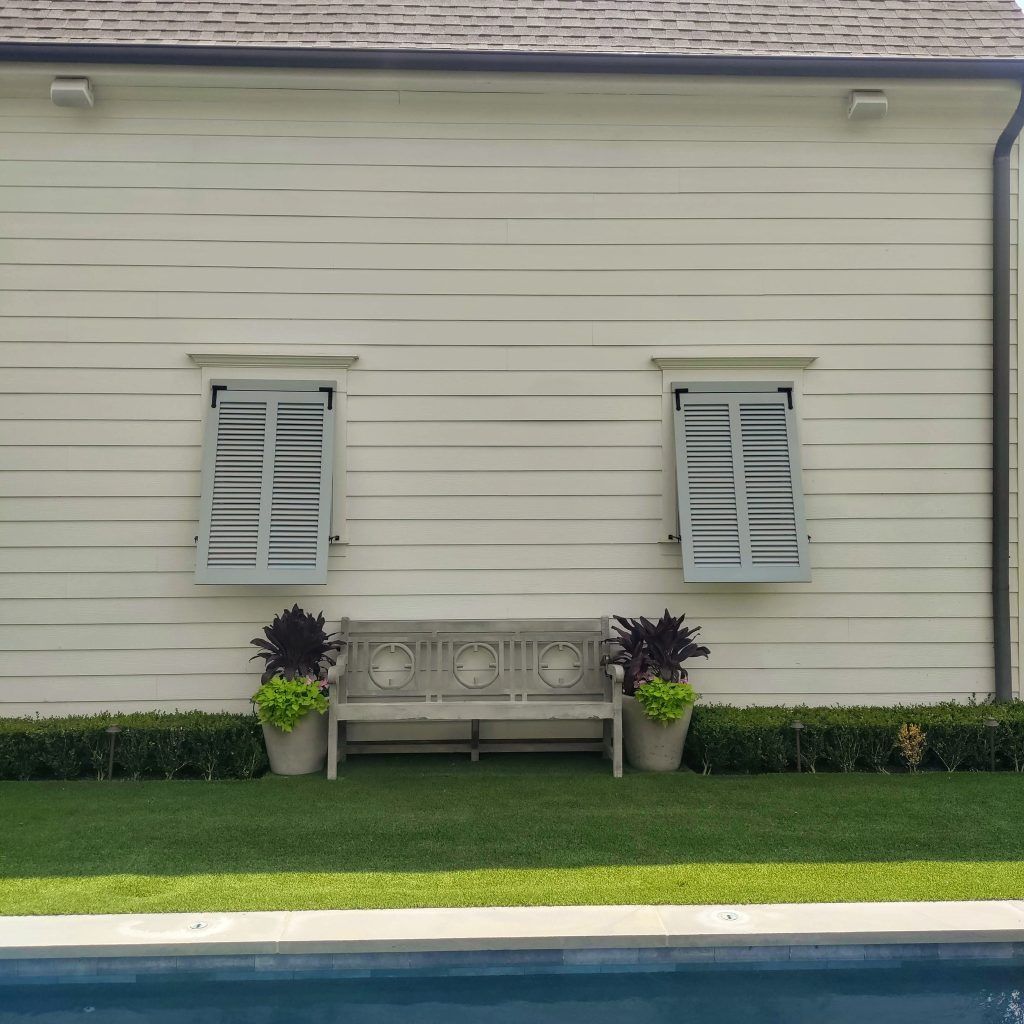 Light-colored house exterior with two shuttered windows, a weathered bench, potted plants, and a swimming pool in the foreground.