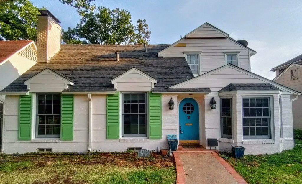 White house with green shutters, blue door, and red brick pathway.