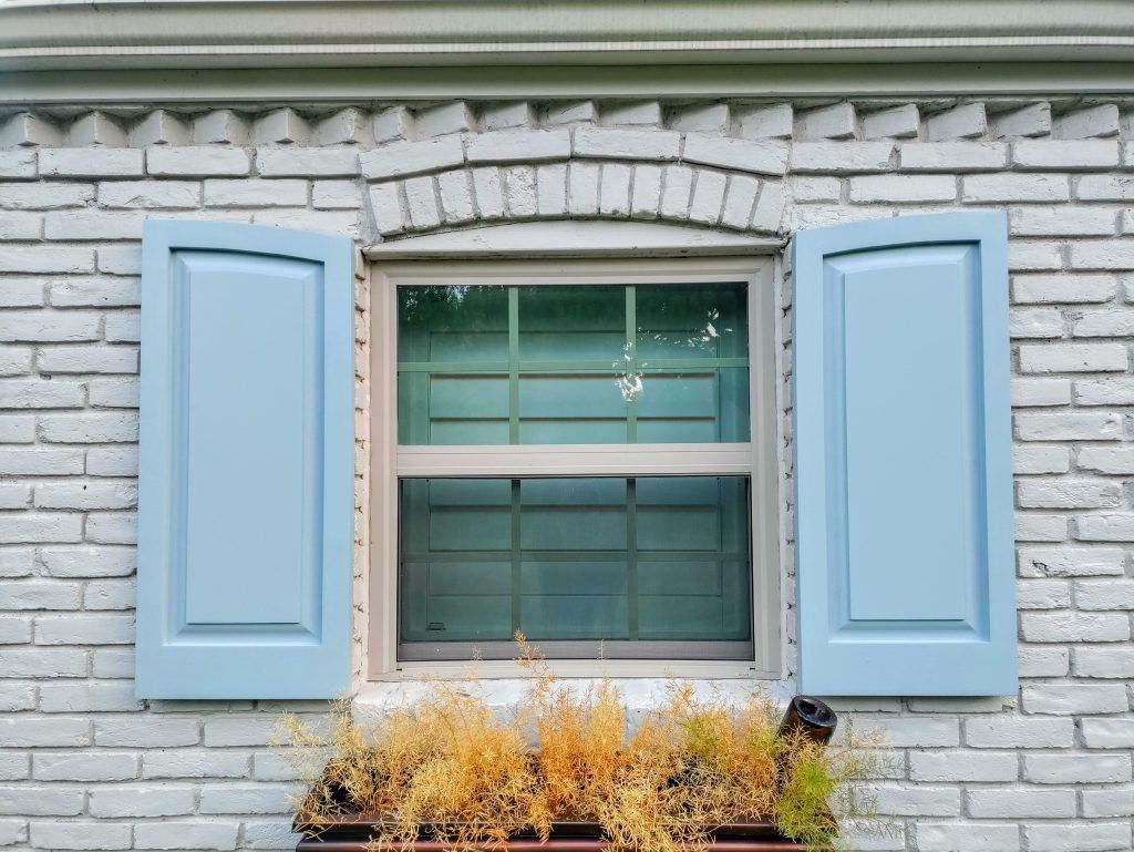 Window with blue shutters and a brick facade, featuring a window box with dead plants.