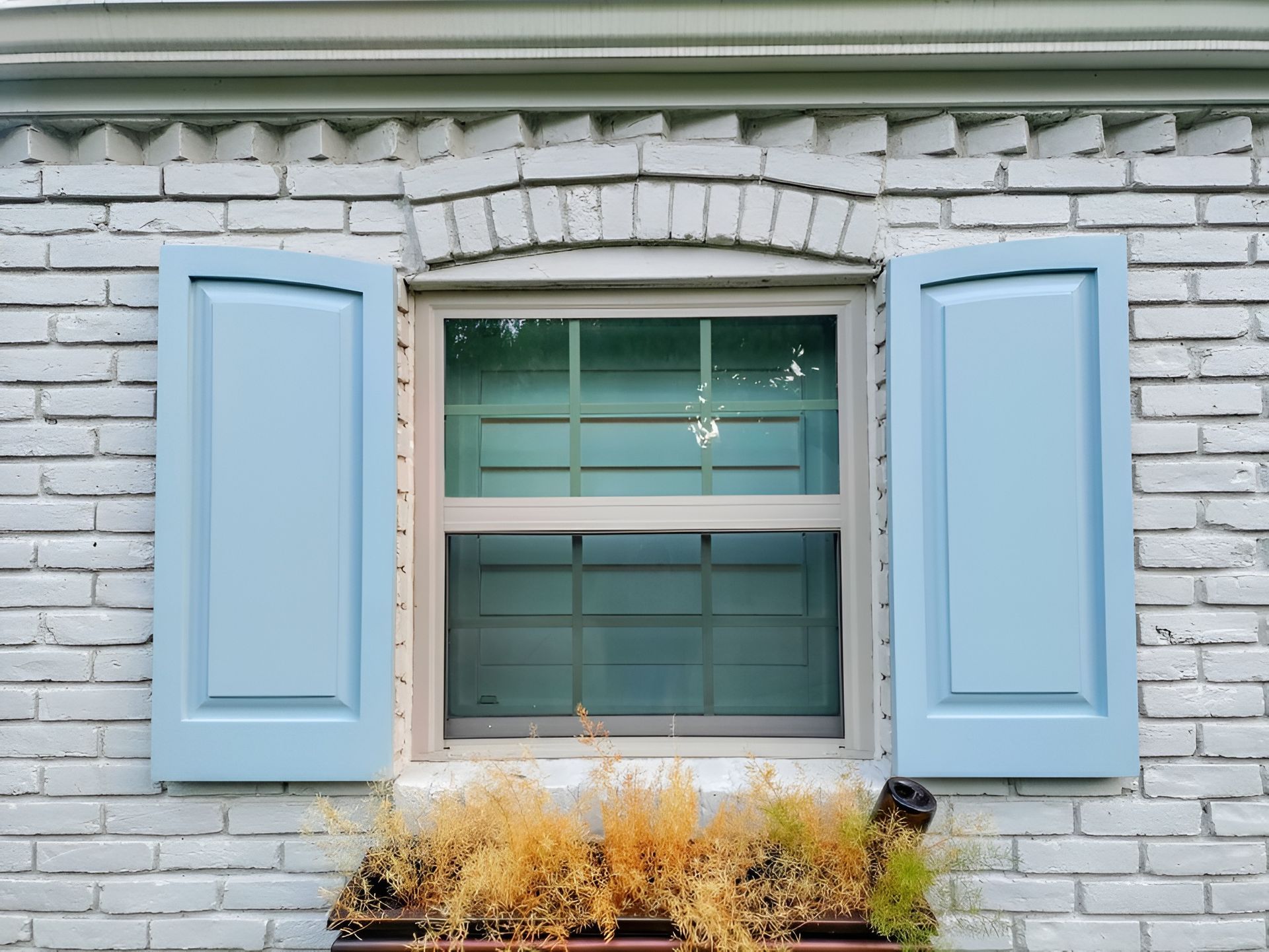 Window with blue shutters on a white brick wall, with a small flower box of dried plants beneath.