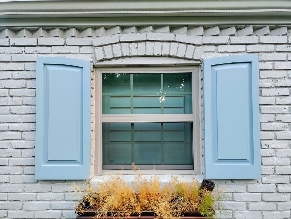 Window with blue shutters on a white brick wall, with a small flower box of dried plants beneath.