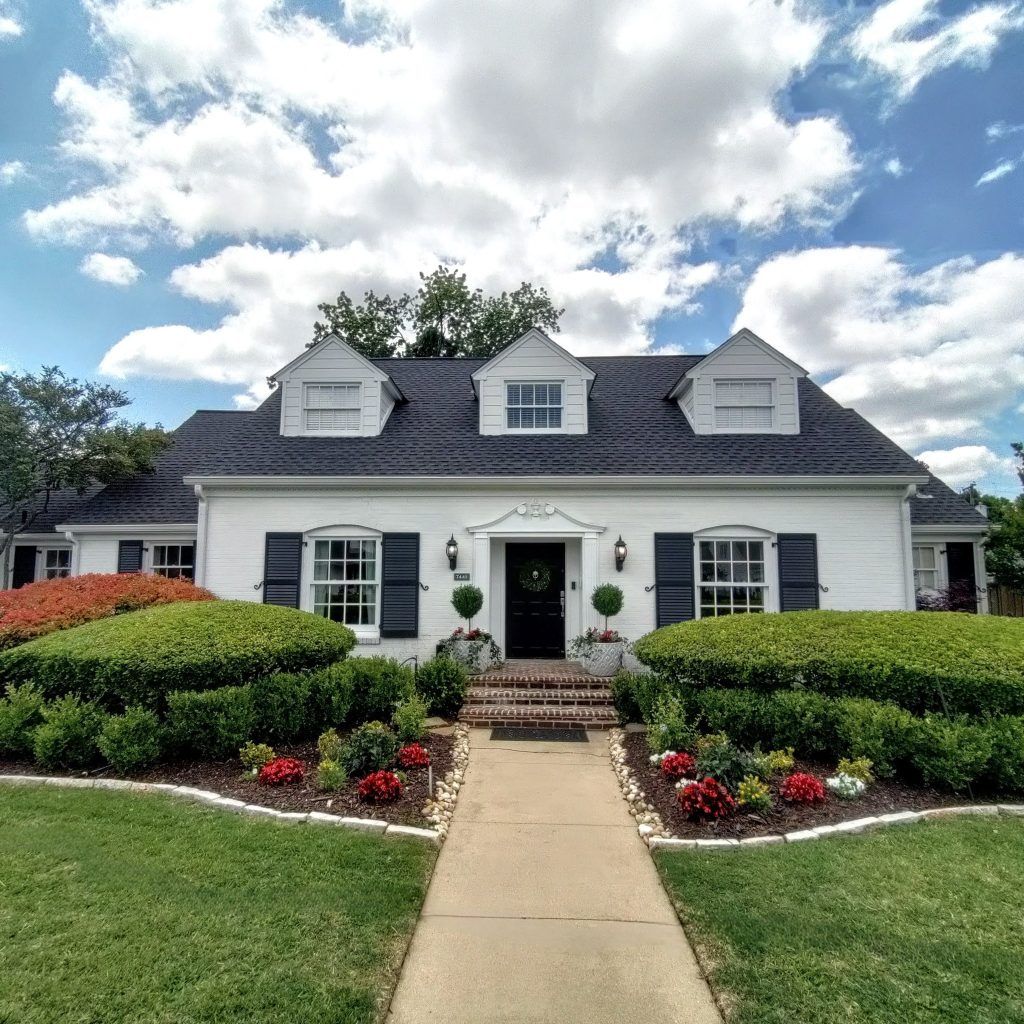 White brick house with black shutters, dormers, and front door. Landscaped front yard and walkway.