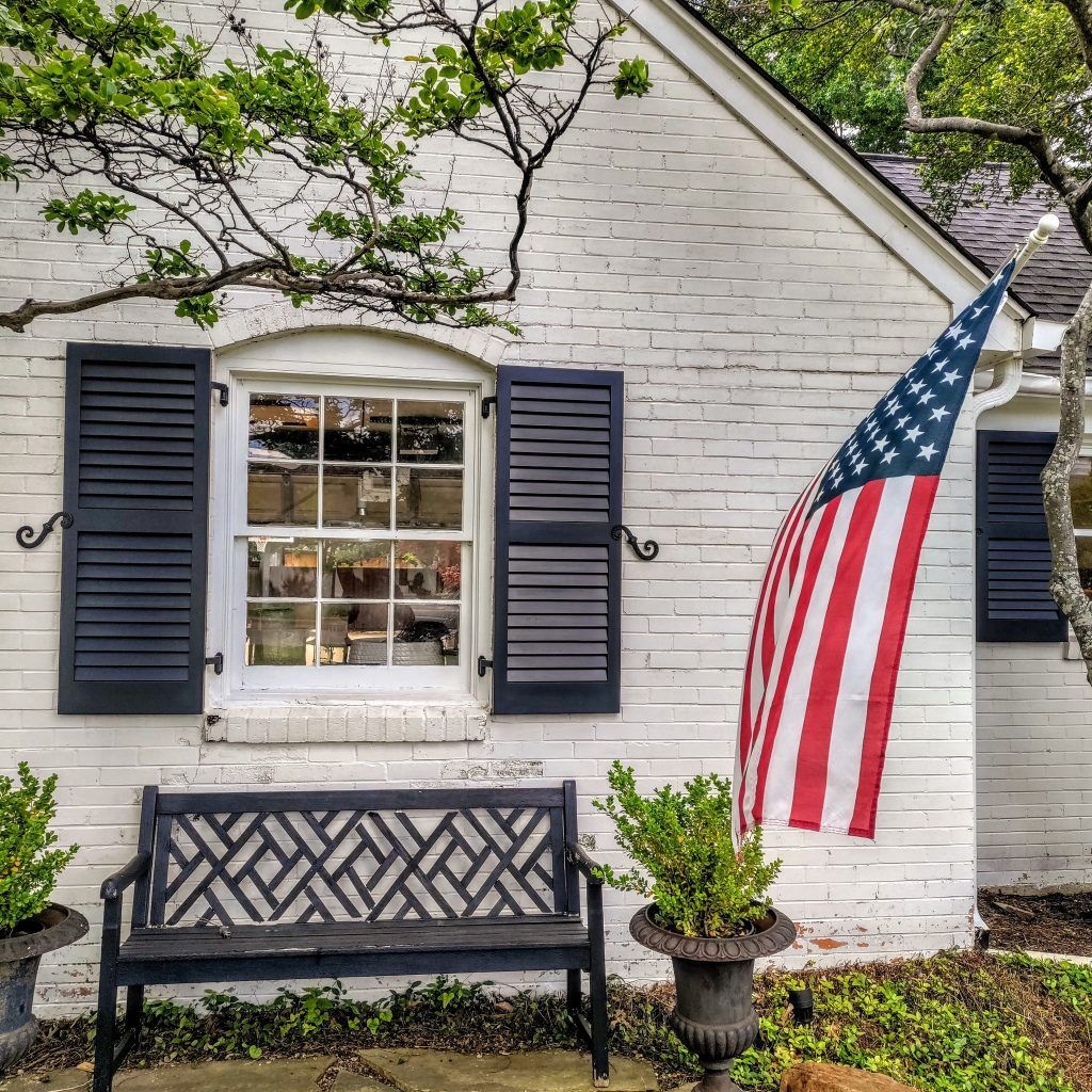 White brick building with black shutters and bench, an American flag, and green plants.