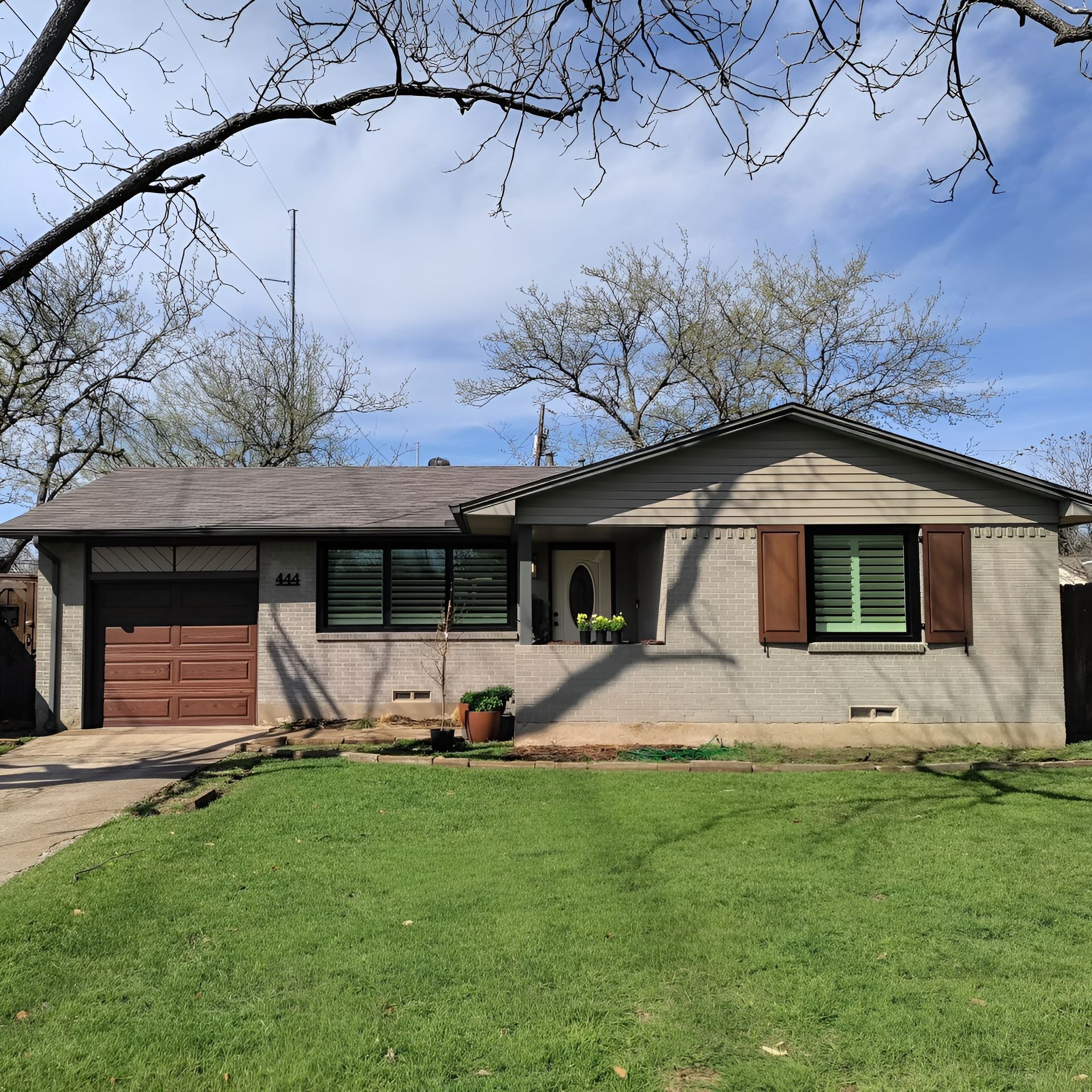 Single-story brick house with brown garage door, shutters, and a front yard with green grass under a blue sky.