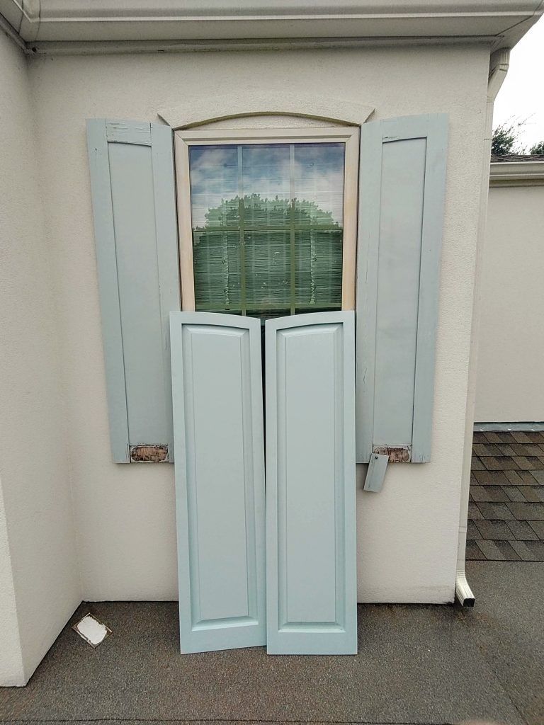 Two light blue shutters lean against a wall below a window with open, matching shutters.