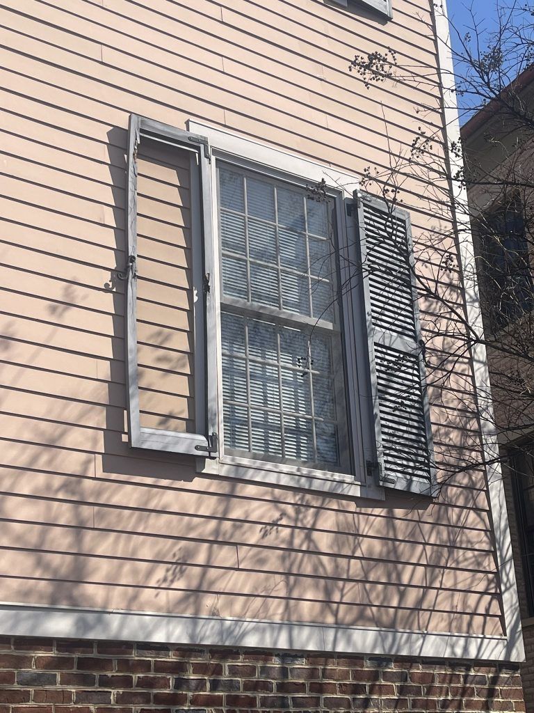 Window with faded shutters on a light pink building. The window is above a brick foundation.