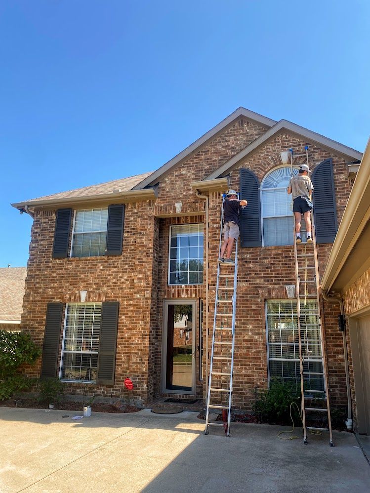 Two people on ladders painting the exterior of a brick house with black shutters, under a clear blue sky.