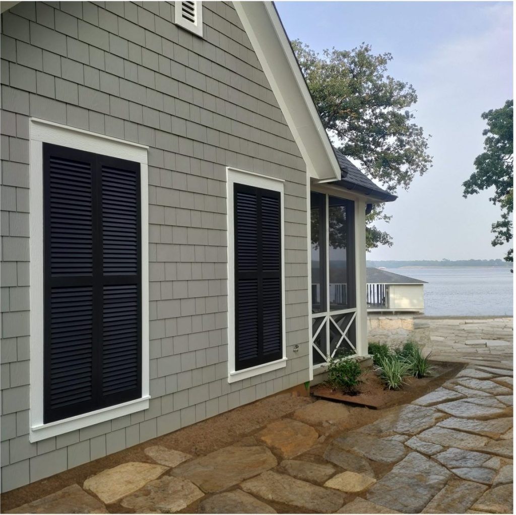Gray house with black shutters, white trim, and stone patio by water.