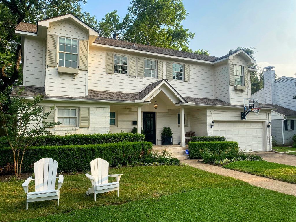 Two-story white house with green lawn. Two white chairs in yard, green hedge in front. Beige shutters.