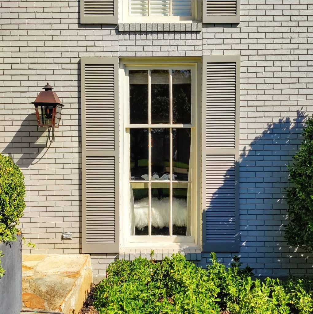 Window with gray shutters on a gray brick building, with a lantern and green bushes.