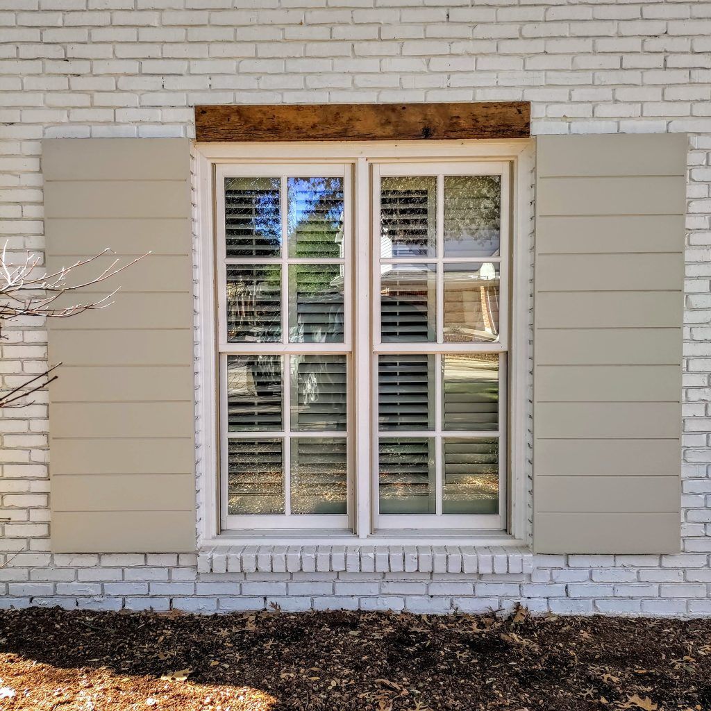 Window with beige shutters on a white brick wall, topped with a dark wood header.