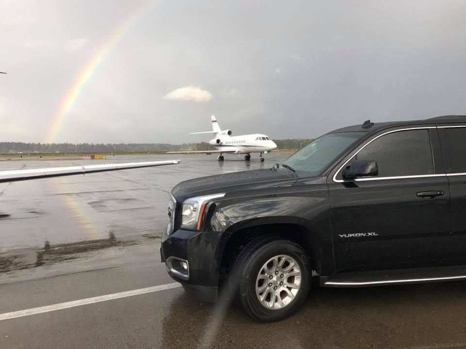Black SUV on an airport tarmac with a private jet and a rainbow in the background under a cloudy sky.