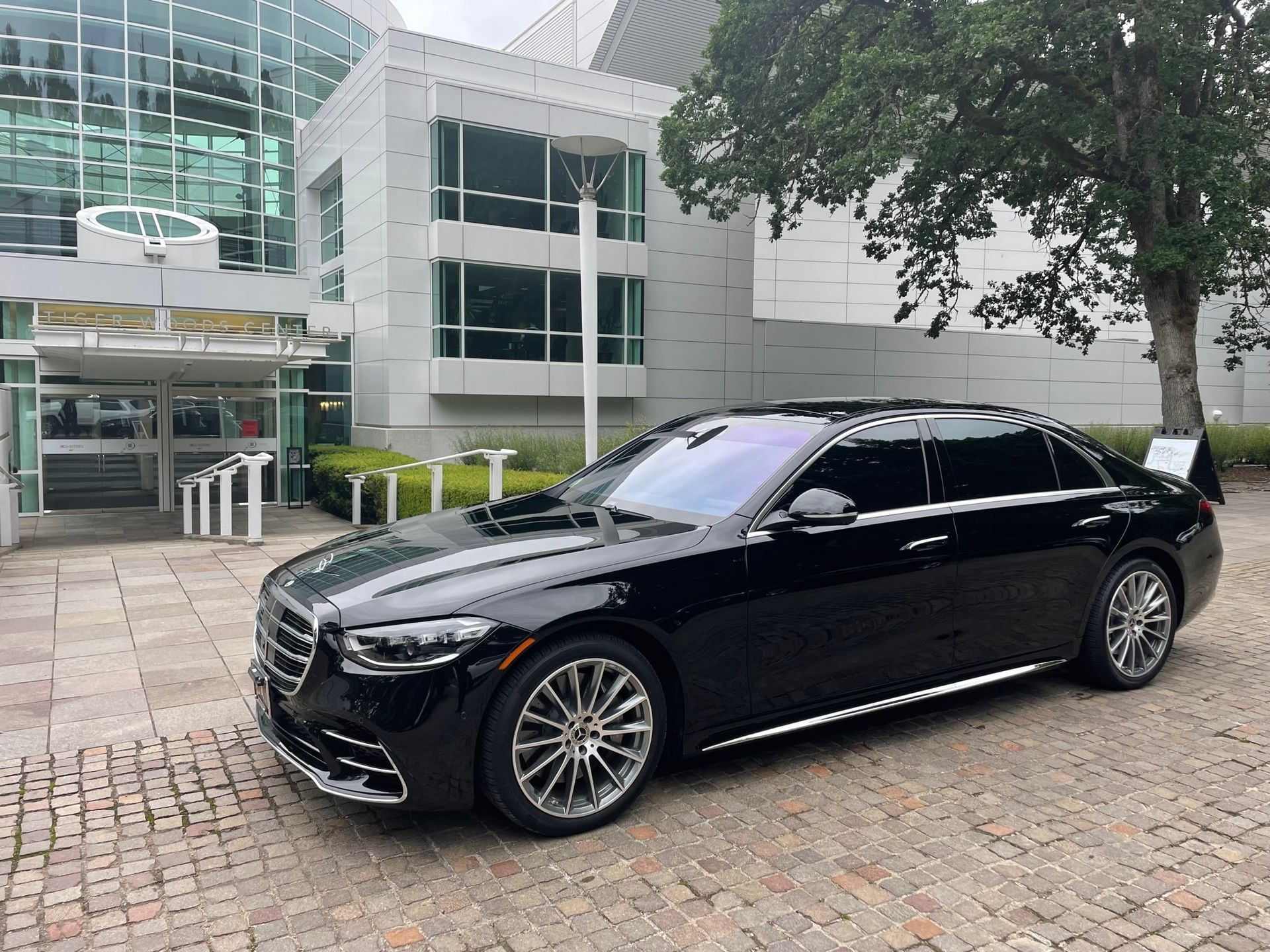 Black Mercedes sedan parked in front of a modern building with a glass facade. The car is on a brick paved area, under a tree.