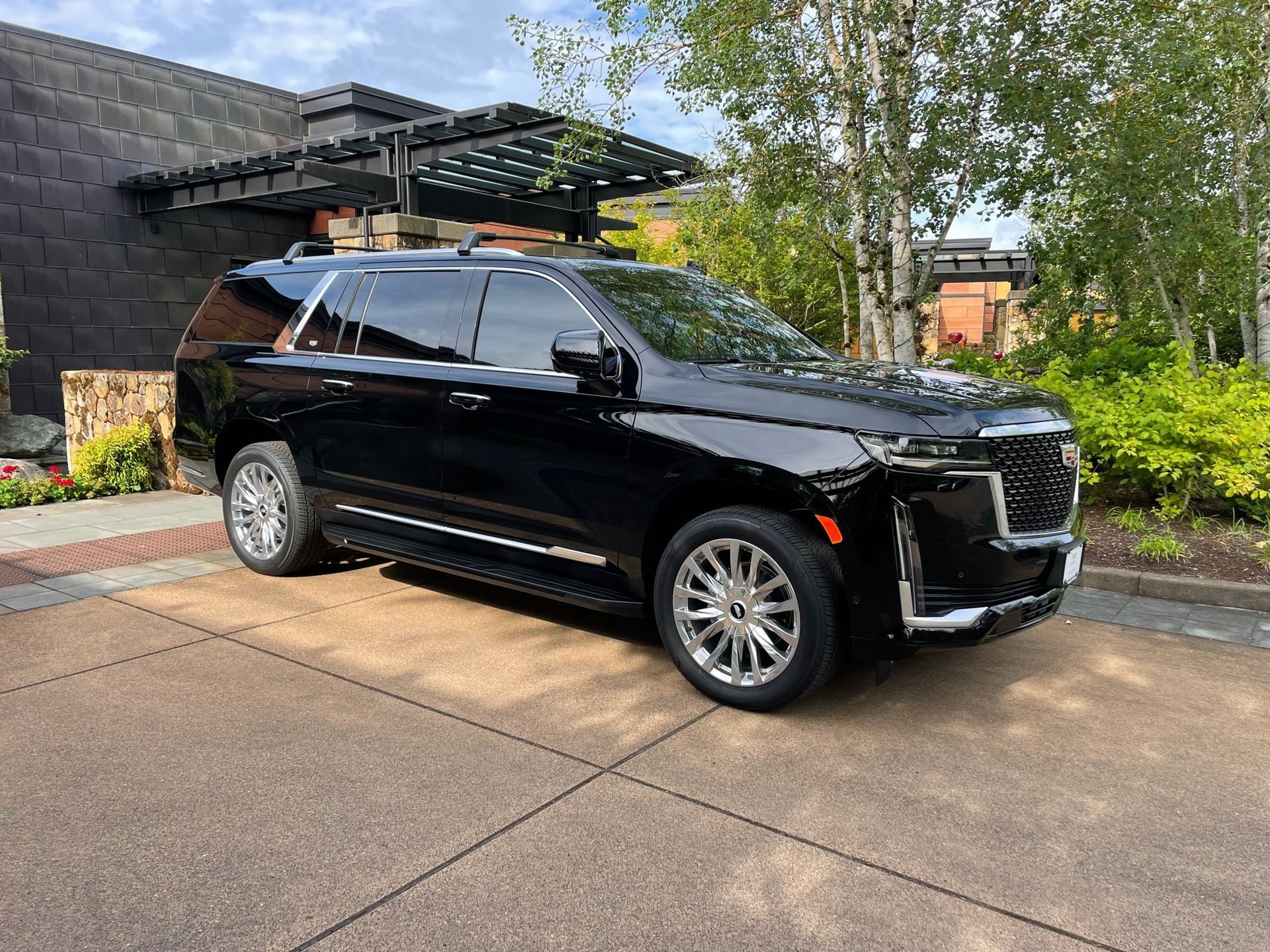 Black Cadillac Escalade parked on a brick driveway in front of a building with a shaded entrance and greenery.