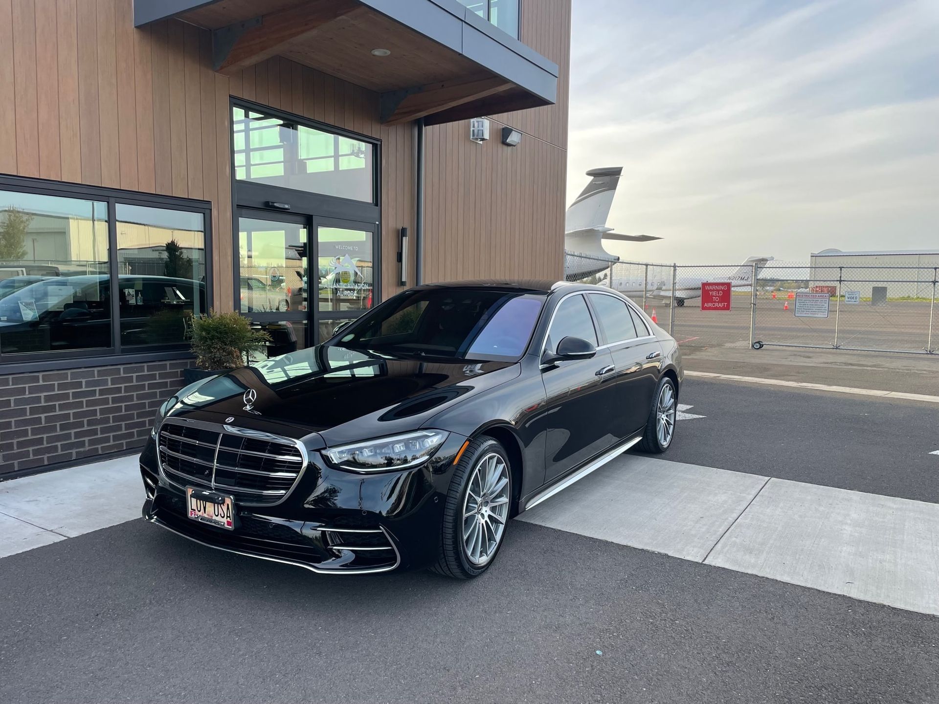 Black Mercedes-Benz sedan parked in front of a building with a view of a plane on a tarmac.