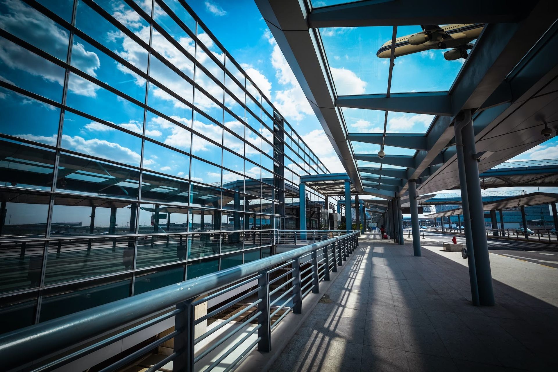 A modern walkway with reflective glass windows and a clear blue sky, likely near an airport or water.
