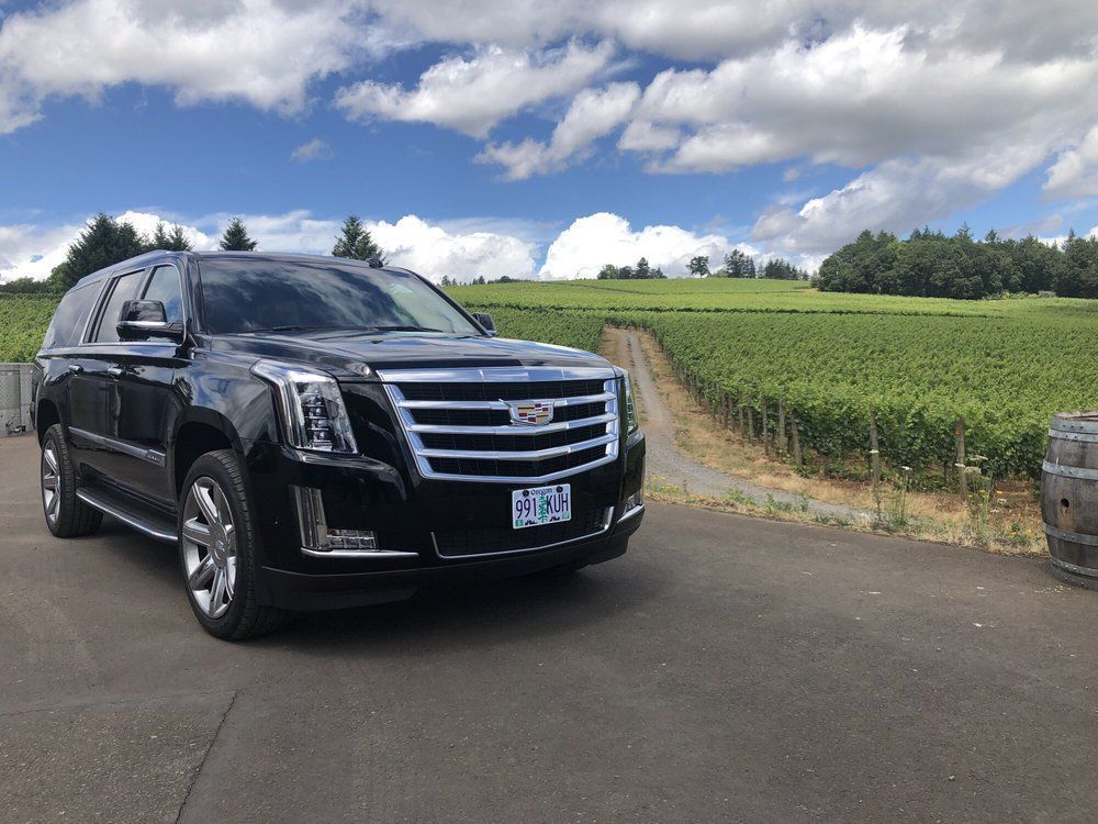 Black Cadillac SUV parked on a paved road in front of a vineyard under a partly cloudy sky.