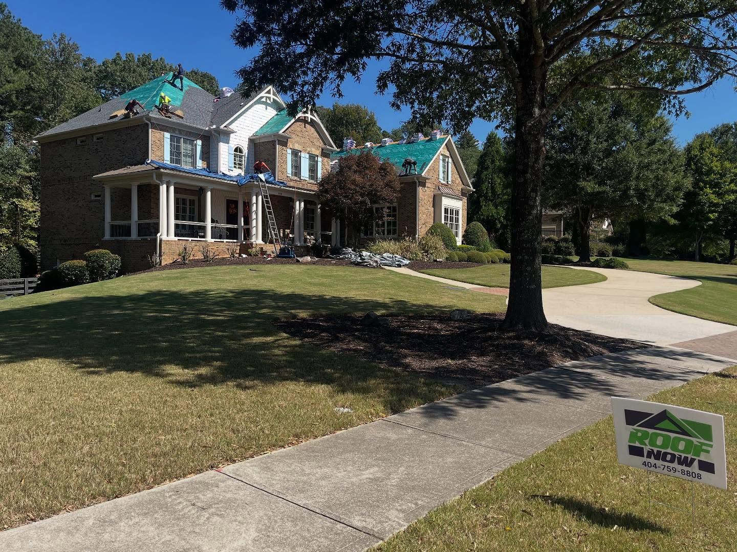 A two-story brick home under renovation with workers on the roof, featuring a sign for a roofing company in the yard.