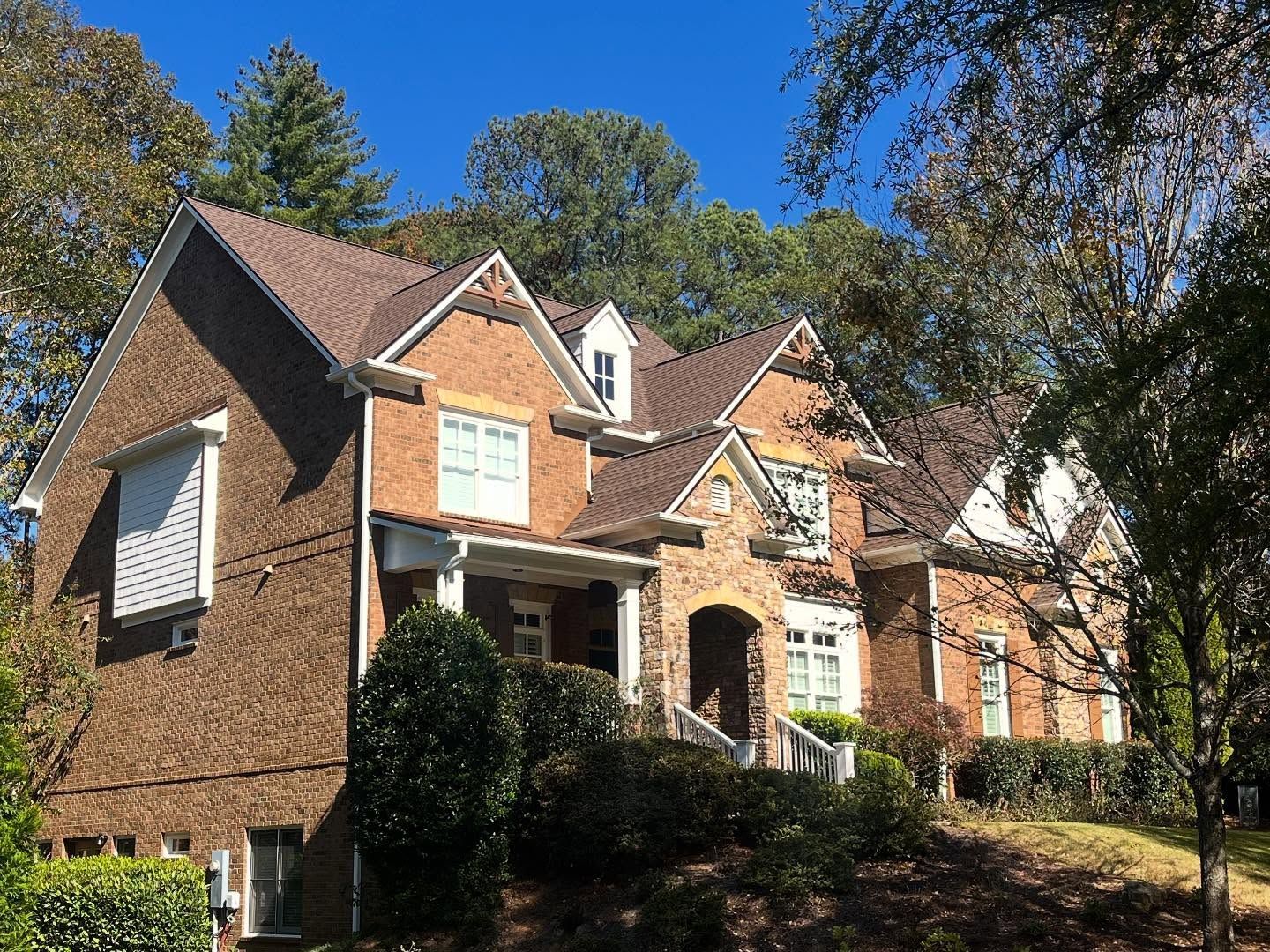 A multi-story brick house with a complex roofline, dormer windows, and a covered entryway, surrounded by trees.