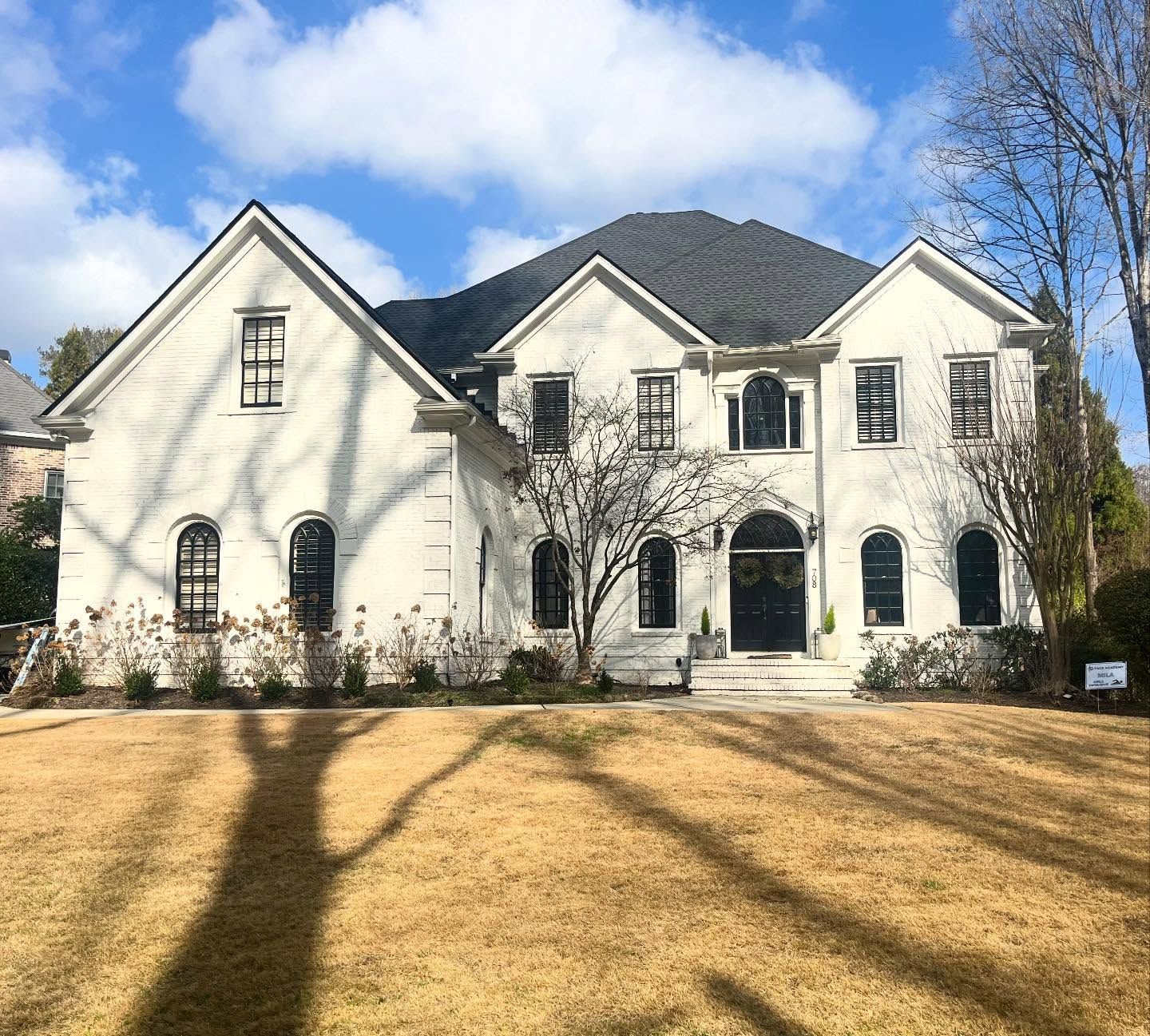 A large, two-story white brick house with a dark roof and black window frames stands behind a dormant, tan-colored lawn.