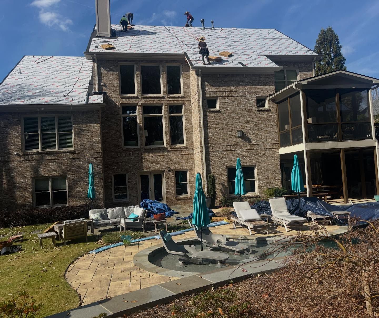 Workers on the roof of a large brick house with a backyard patio featuring lounge chairs, a fire pit, and a screened porch.