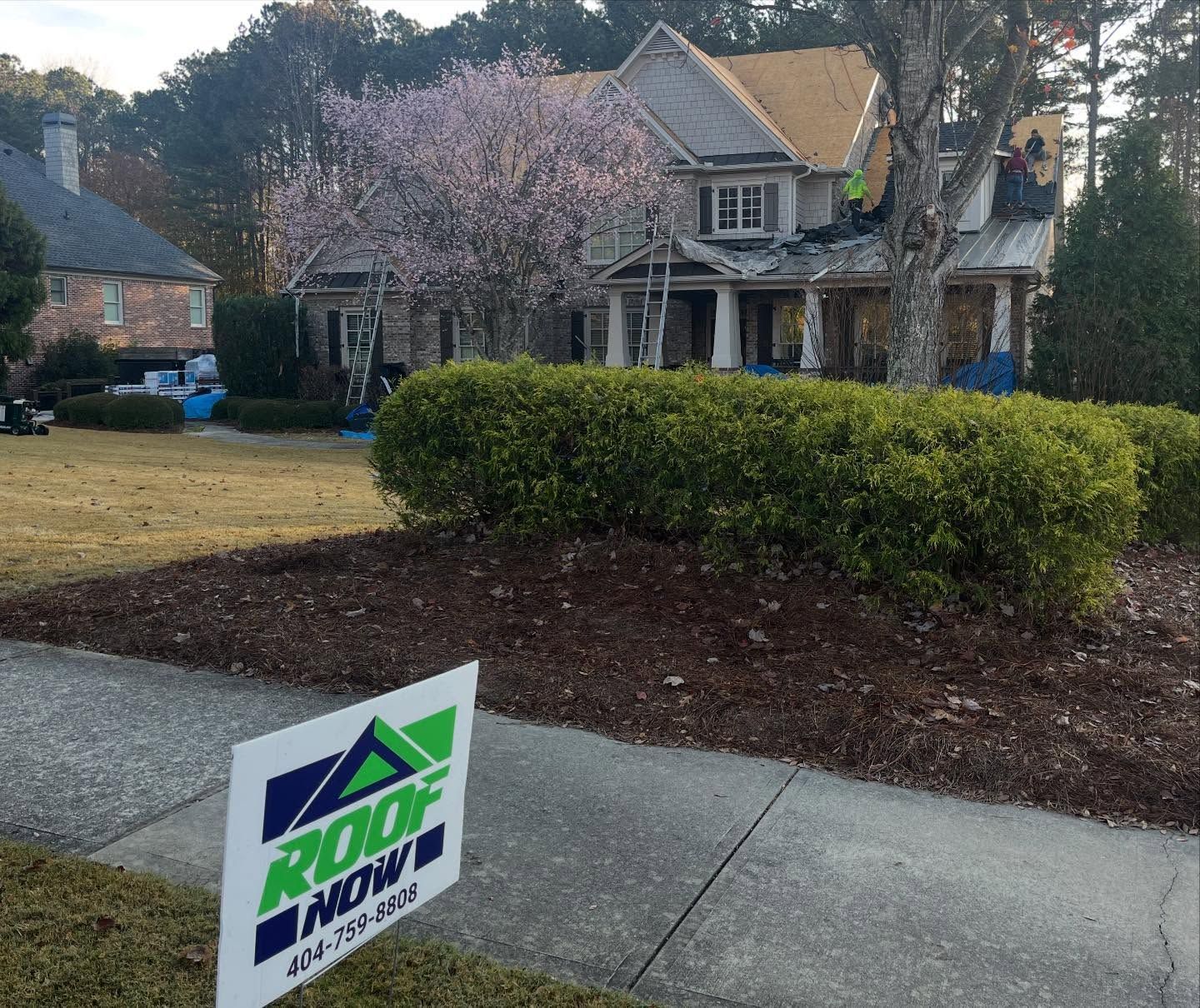 A Roof Now sign stands on a lawn in front of a residential house undergoing roof repairs, featuring a blooming pink tree.
