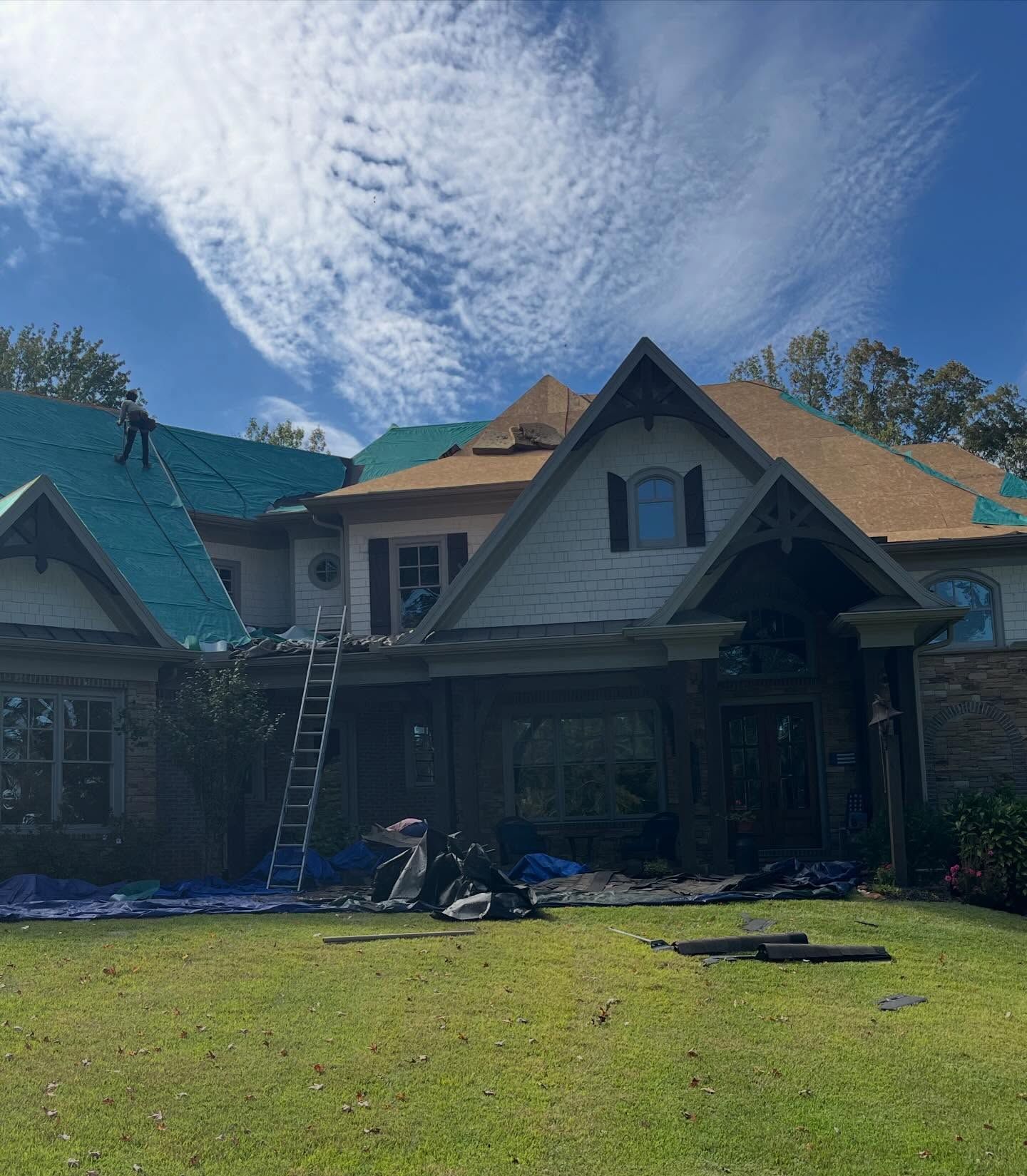 Workers installing a new roof on a large house with blue underlayment and brown shingles under a sunny sky.