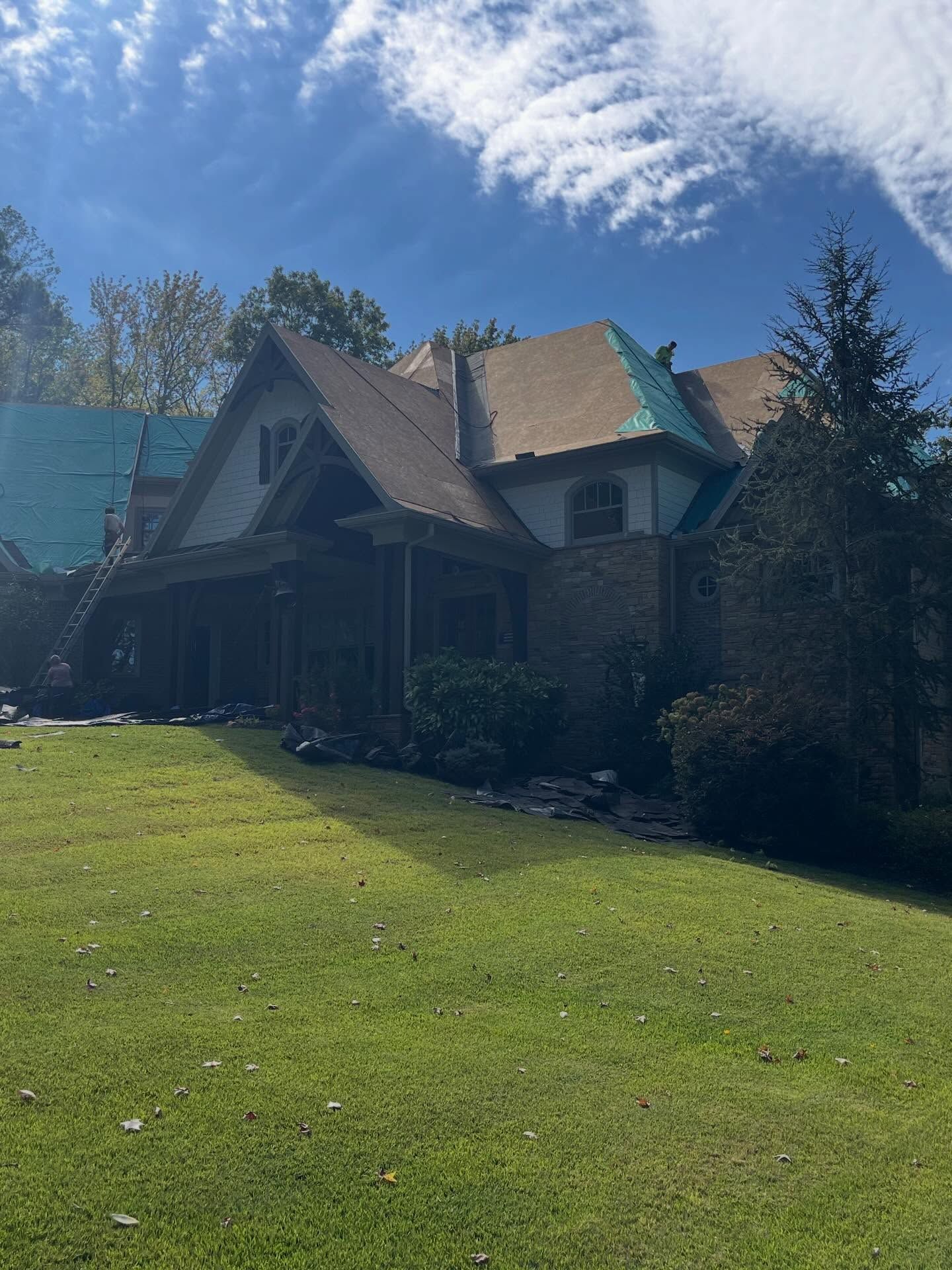 A stone-and-stucco house with a partially replaced roof under a bright blue sky, with an evergreen tree on the right.