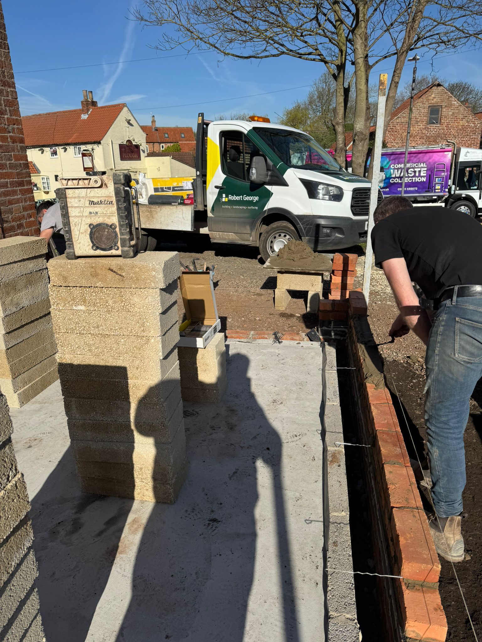 Construction worker laying bricks; truck and building materials on a sunny day.