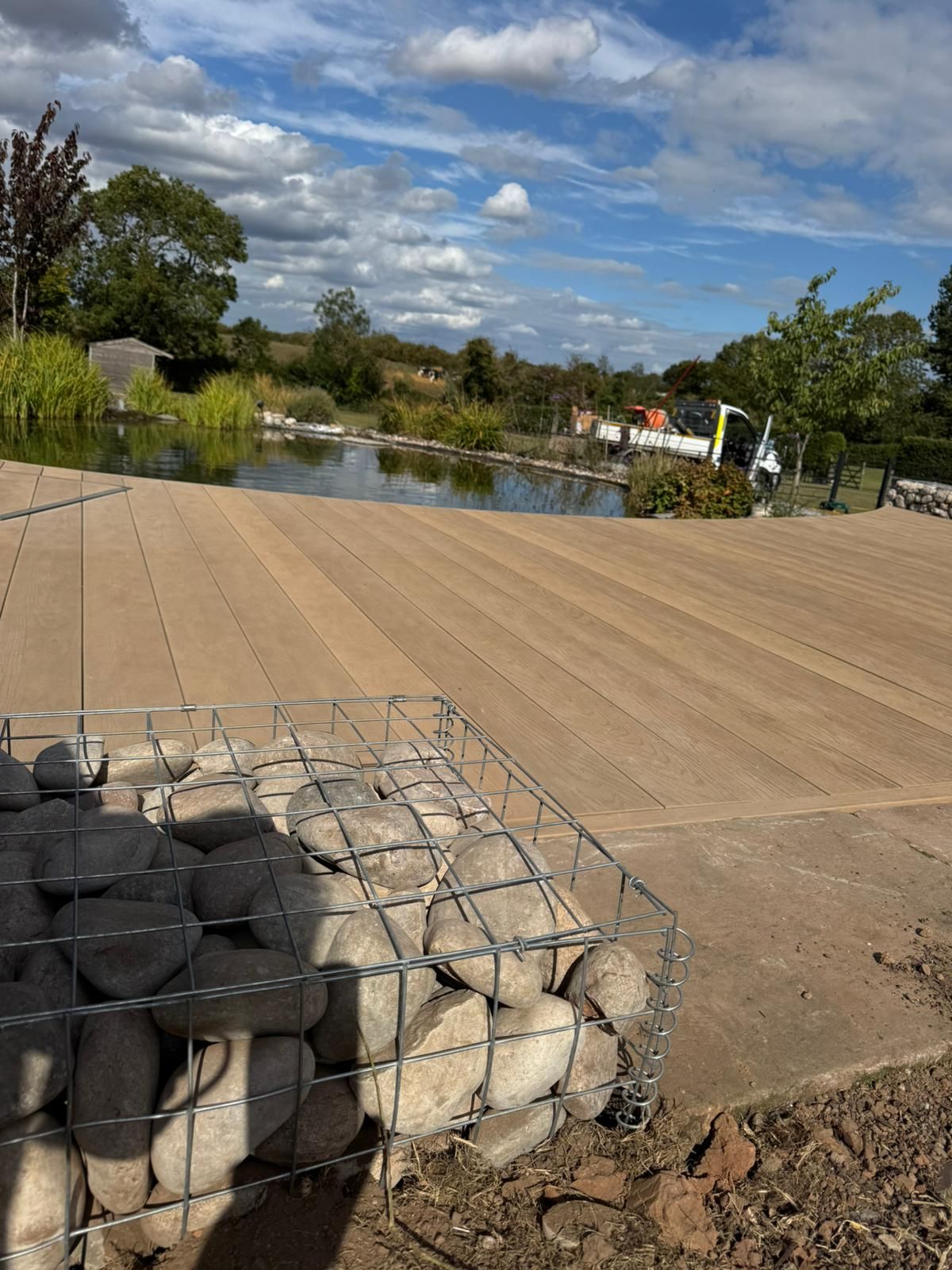 Stone-filled wire cage in the foreground, with a wood deck and pond in the distance under a partly cloudy sky.