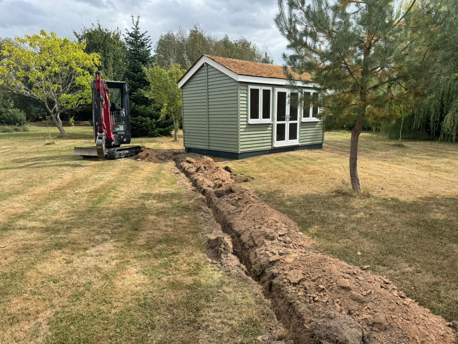 A mini excavator digging a trench in a grassy yard, near a green shed with white trim.
