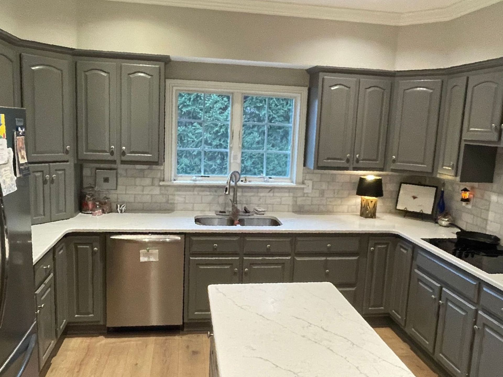 A kitchen with gray cabinets , white counter tops , stainless steel appliances and a large island.