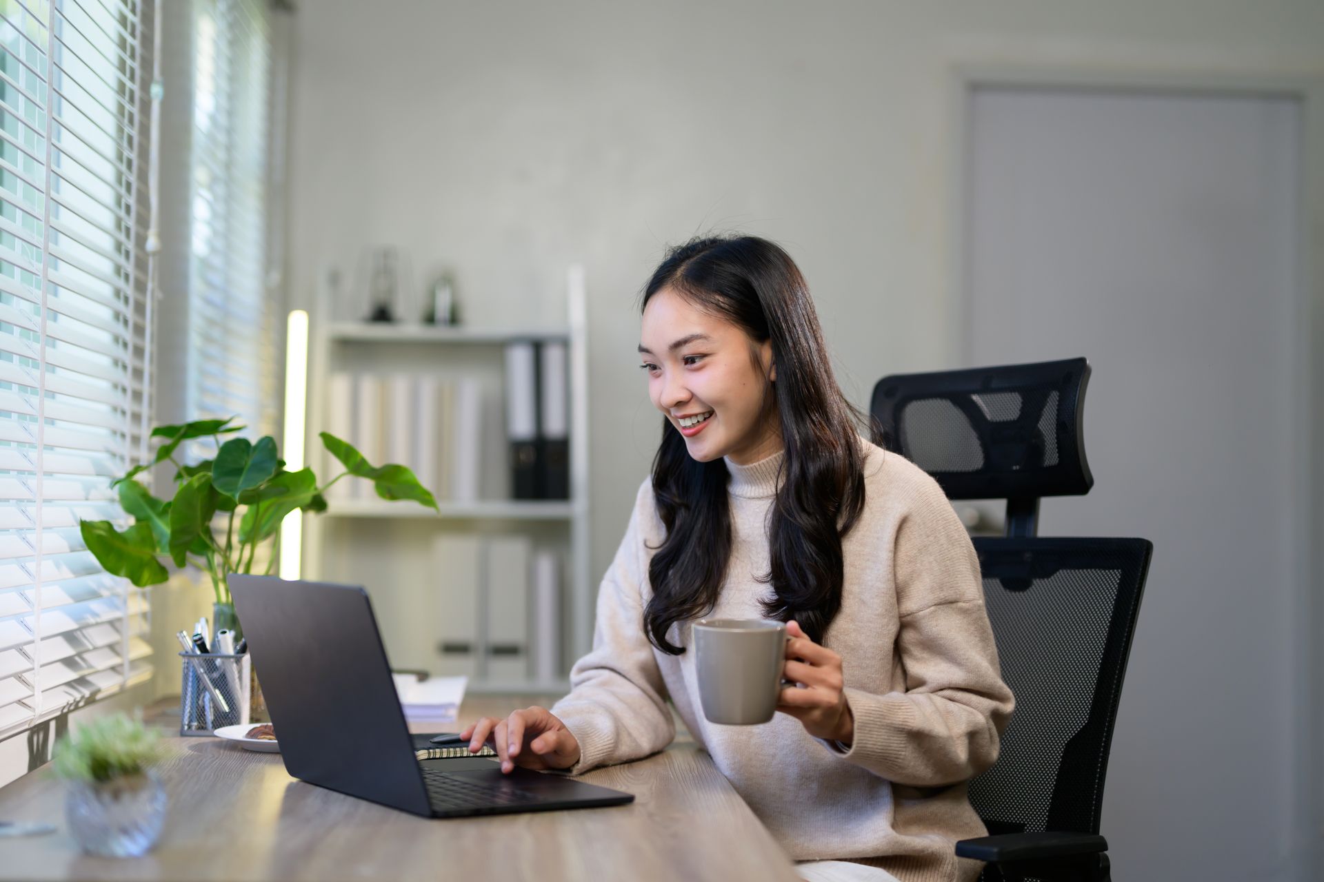 Woman at desk with laptop, smiling, holding mug, near window with blinds.
