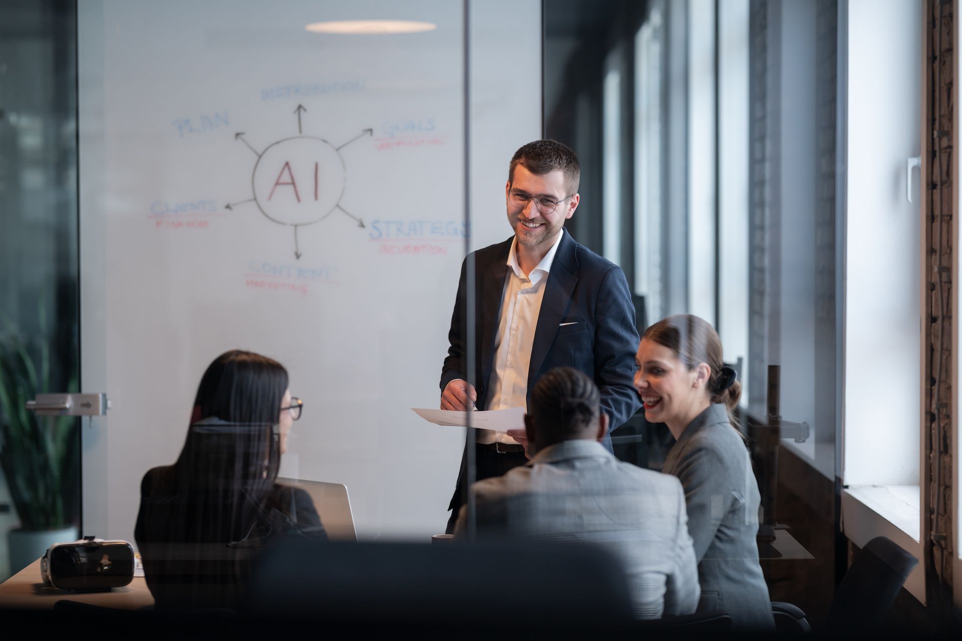 Business meeting in a glass-walled conference room, with a presenter speaking beside a whiteboard labeled “AI.”