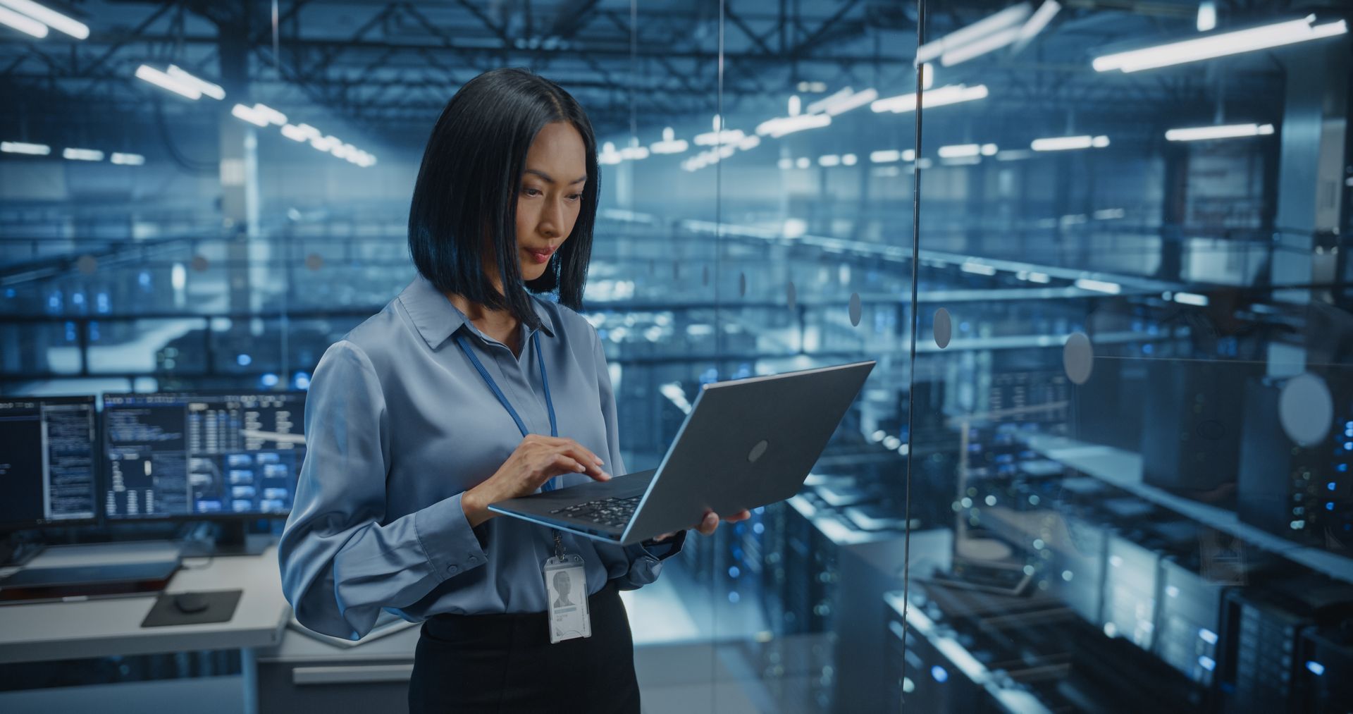 Woman in a blue shirt using a laptop in a data center, rows of servers visible in the background.