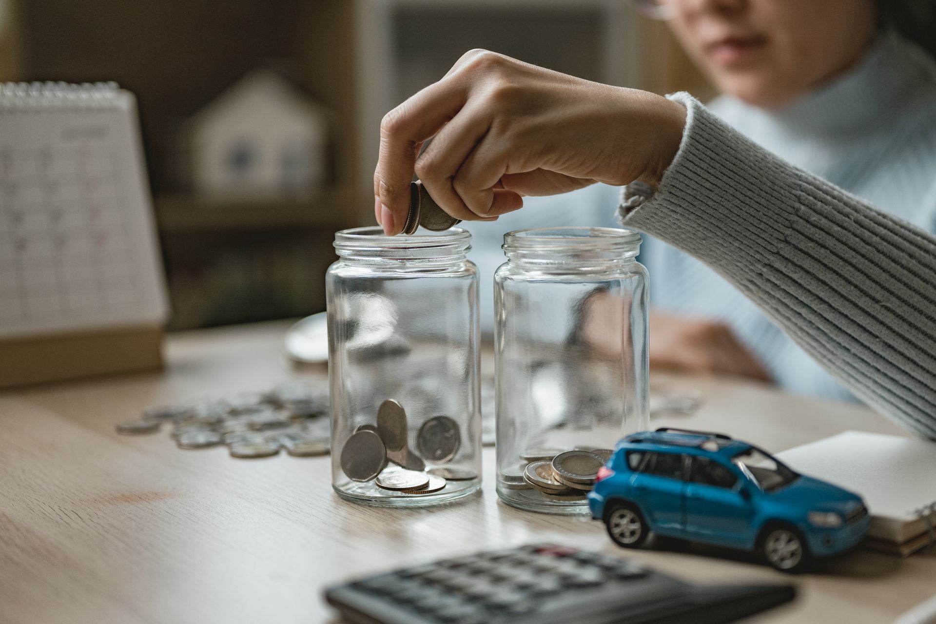 Hand placing coins into glass jars beside a calculator and blue toy car on a desk