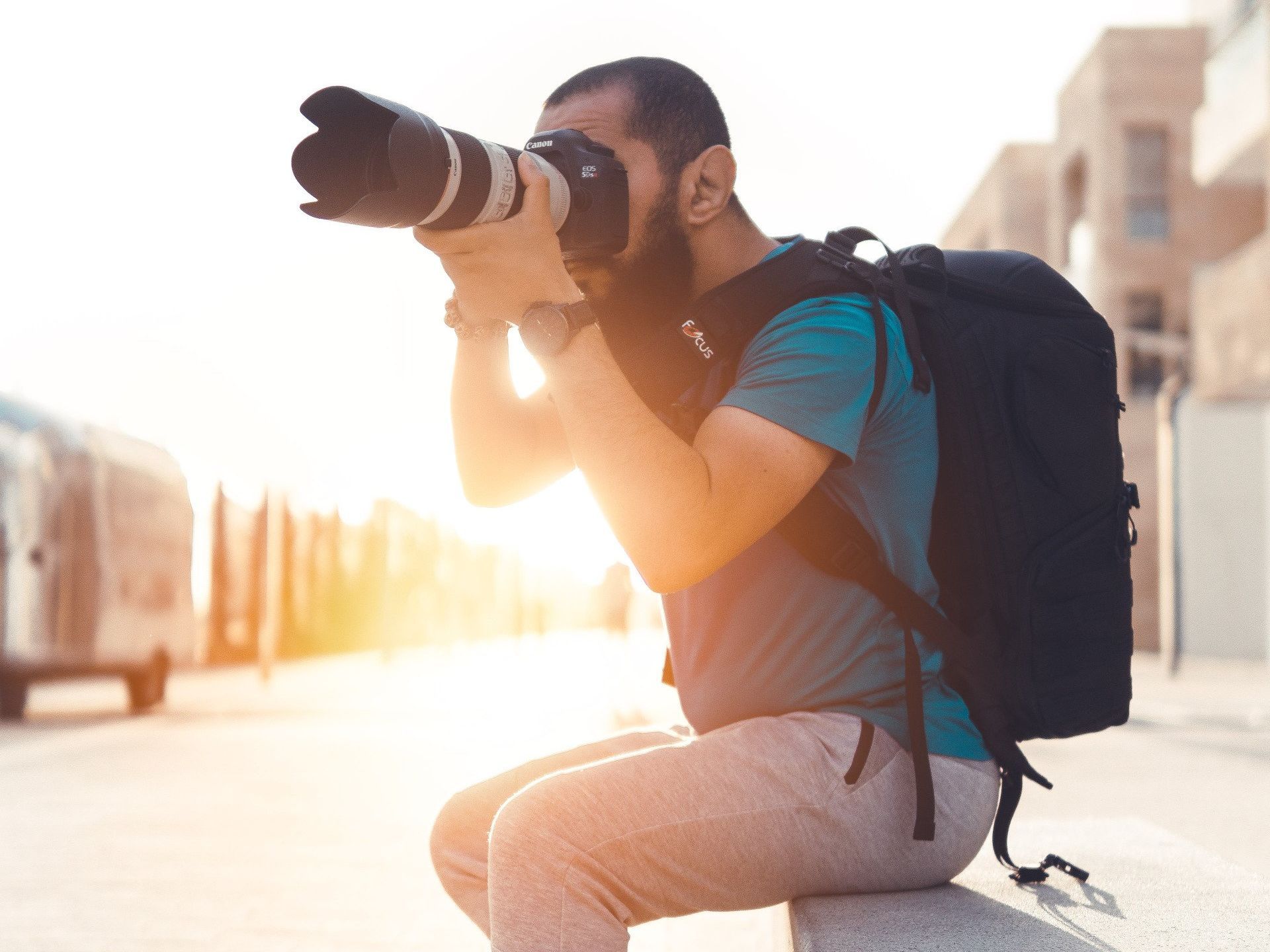 A man with a backpack is kneeling down and taking a picture with a camera.