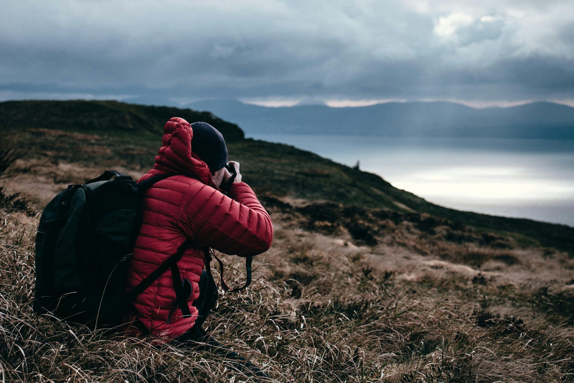 A man in a red jacket is sitting on top of a hill taking a picture.