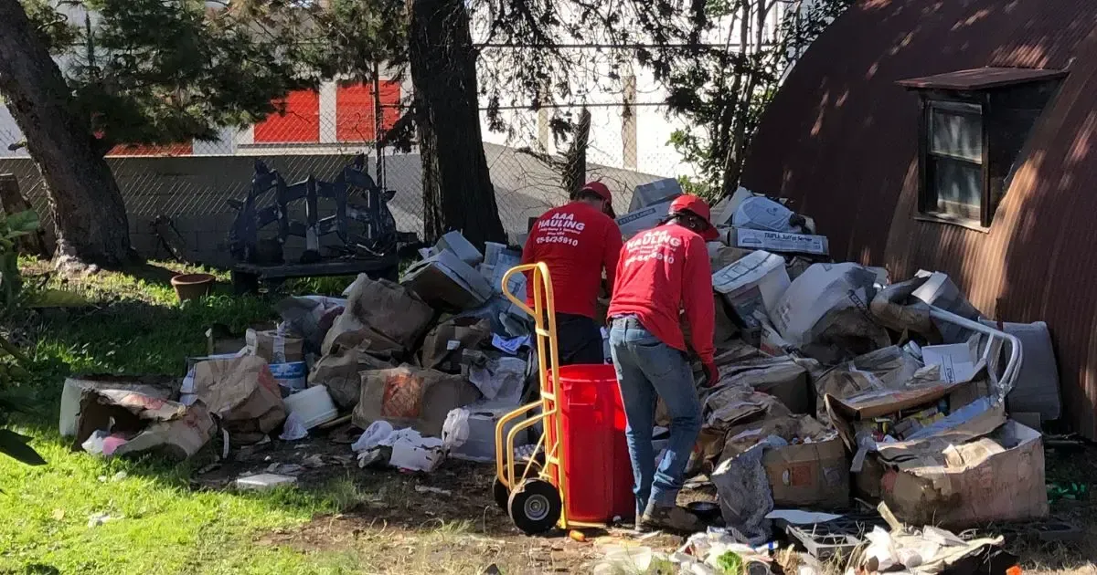 Two people in red shirts sort through trash bags and debris next to a cart.