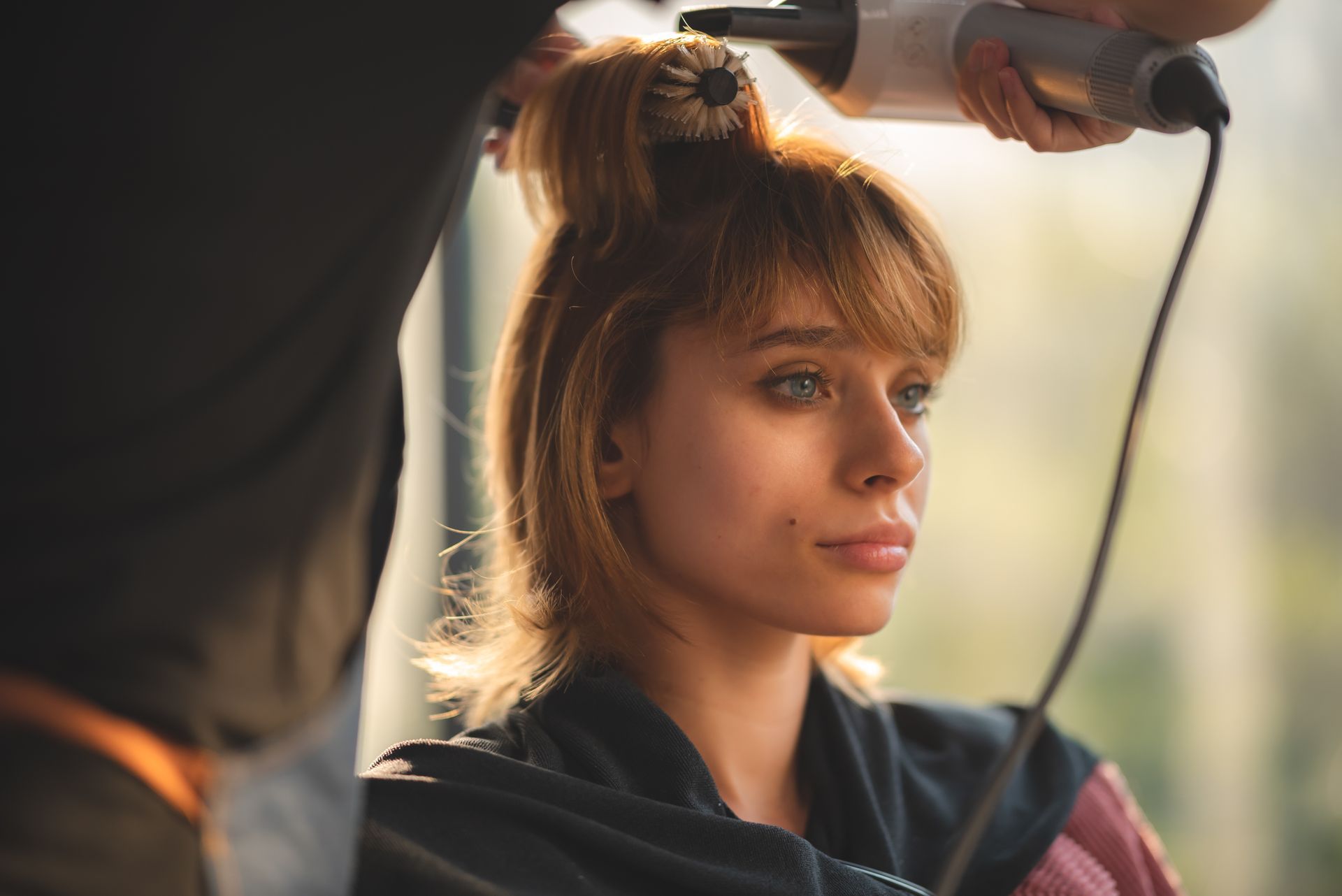 Woman getting her hair styled; stylist using a hair dryer and round brush. Natural light, neutral tones.