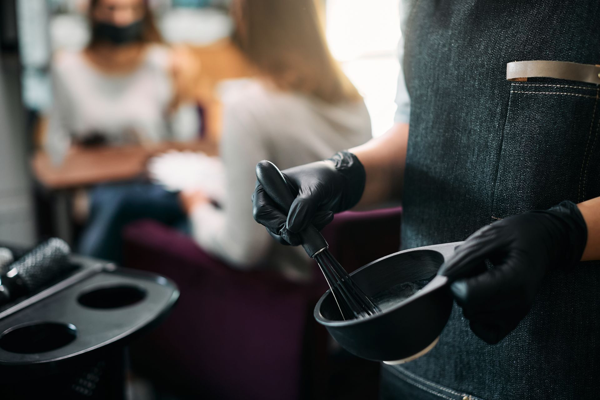 Hair stylist mixes dye in a black bowl, wearing black gloves, in a salon setting with two clients blurred in the background.