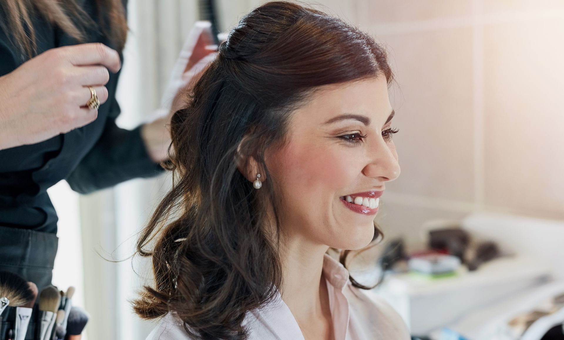 A woman with a half-up hairdo smiles as a stylist does her hair, indoors.