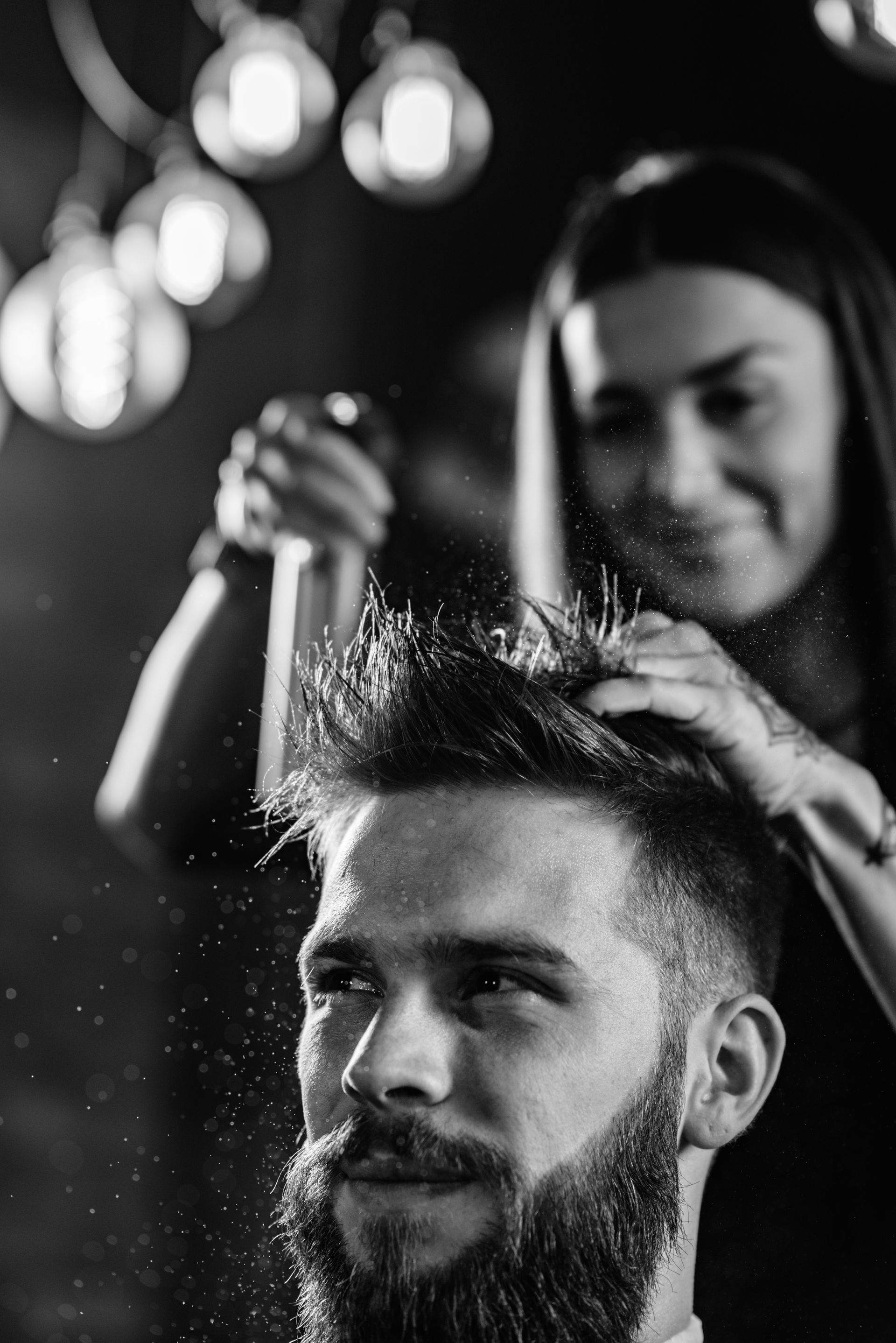 Woman spraying man's hair in a barbershop. Man with beard looks to the side, happy expression. Black and white.
