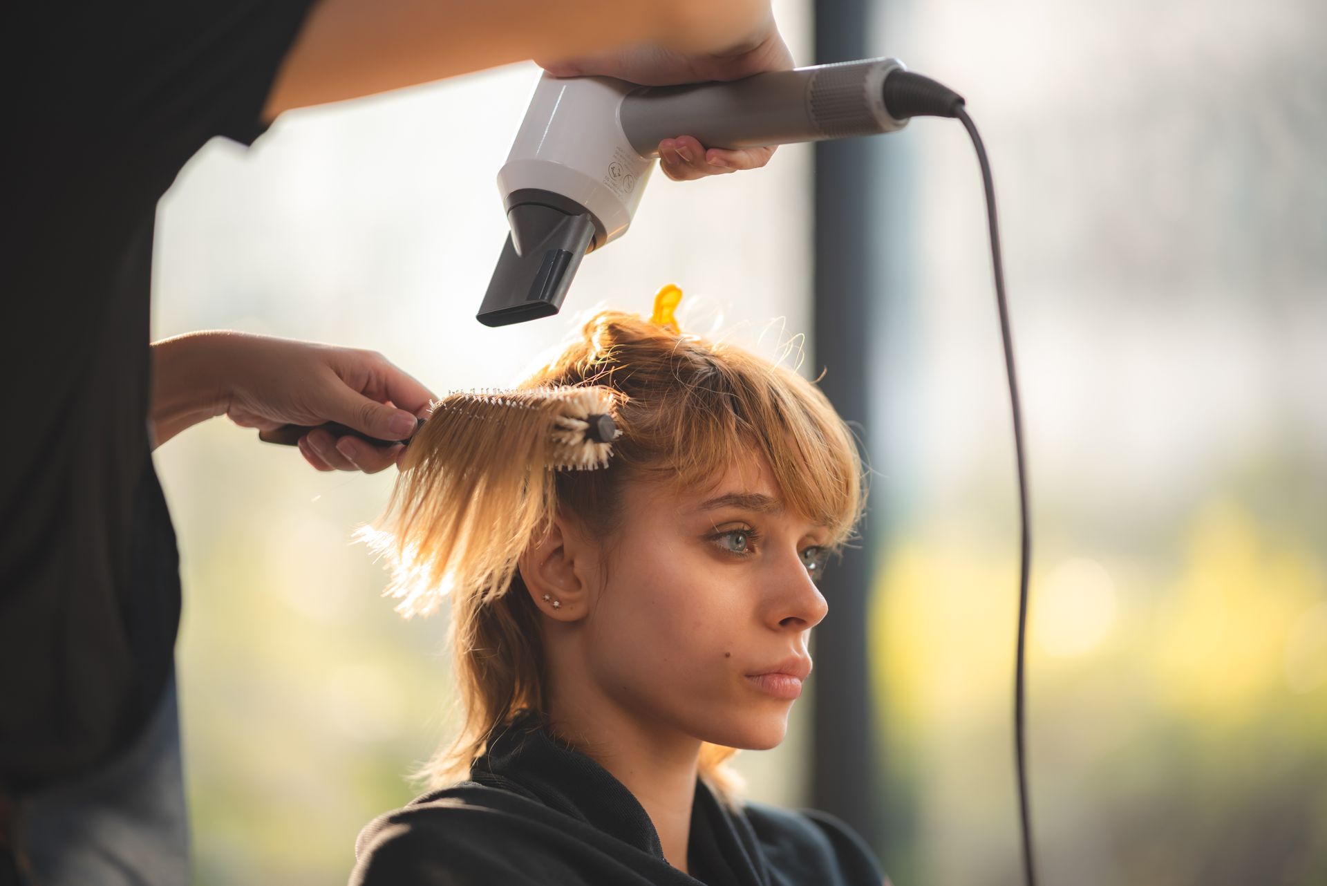A stylist using a hair dryer on a young woman in a salon setting.