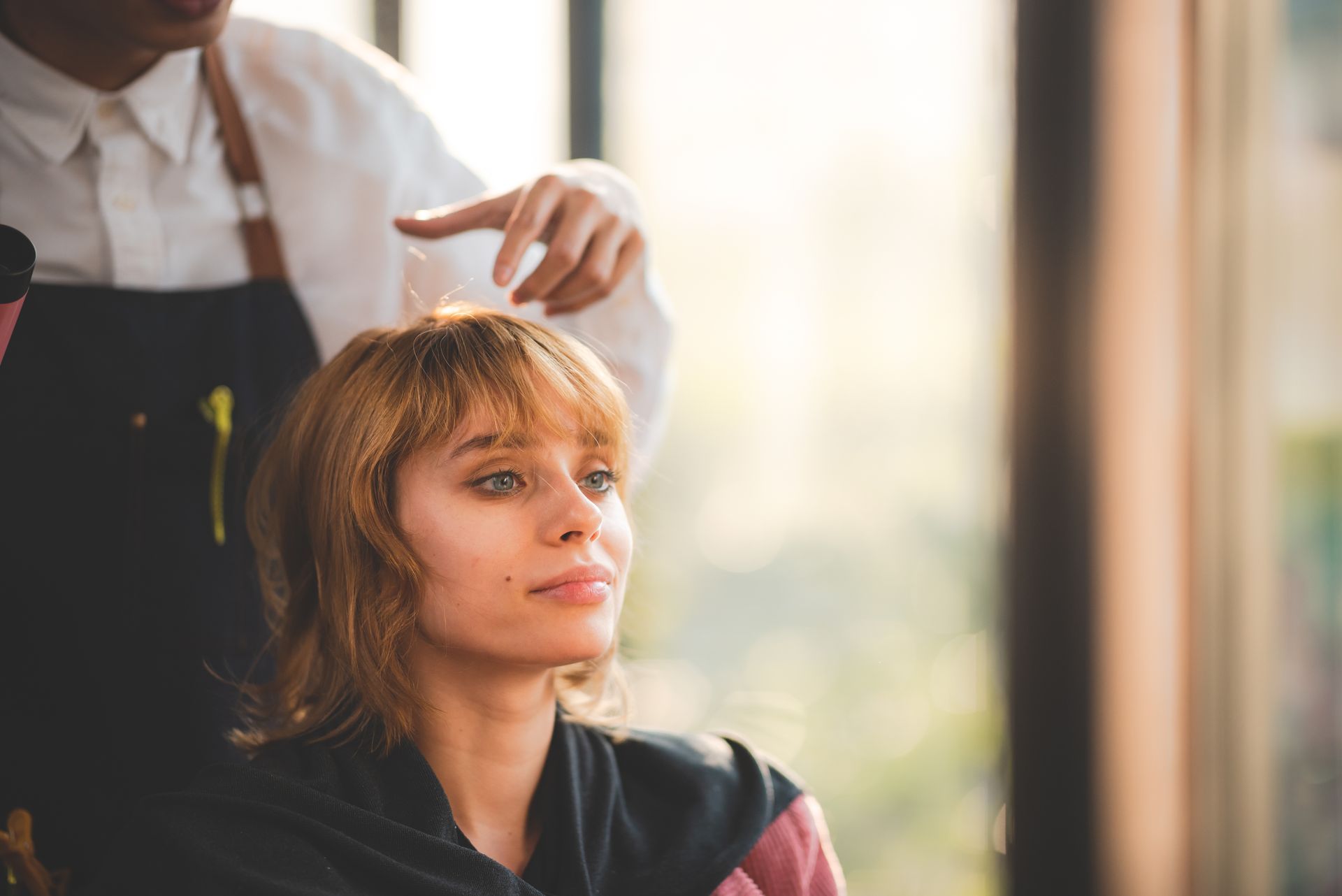 Woman getting hair styled in a salon. Hairdresser's hands are near her head. Warm lighting from a window.