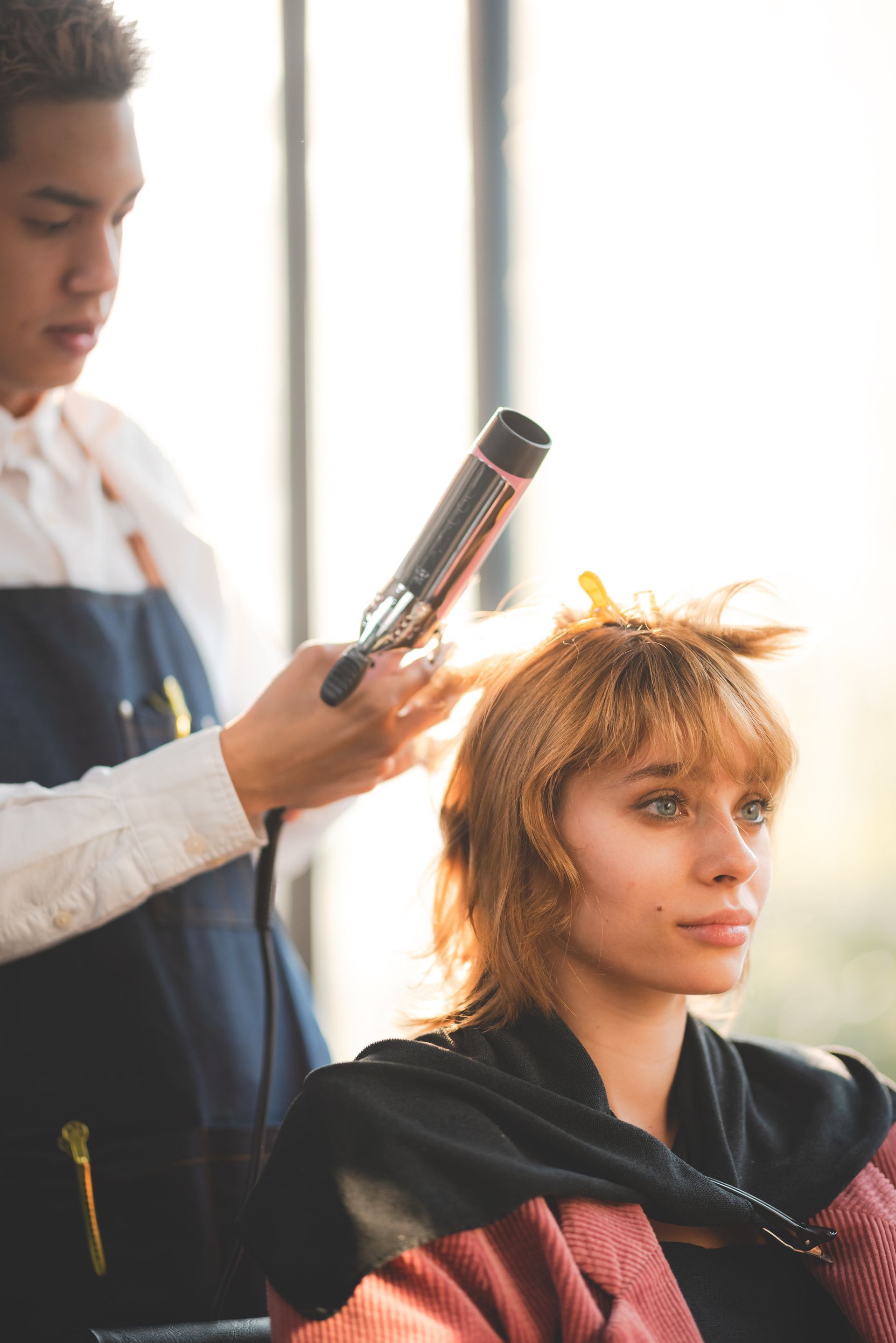Hair stylist curling a woman's orange hair in a salon; warm lighting.