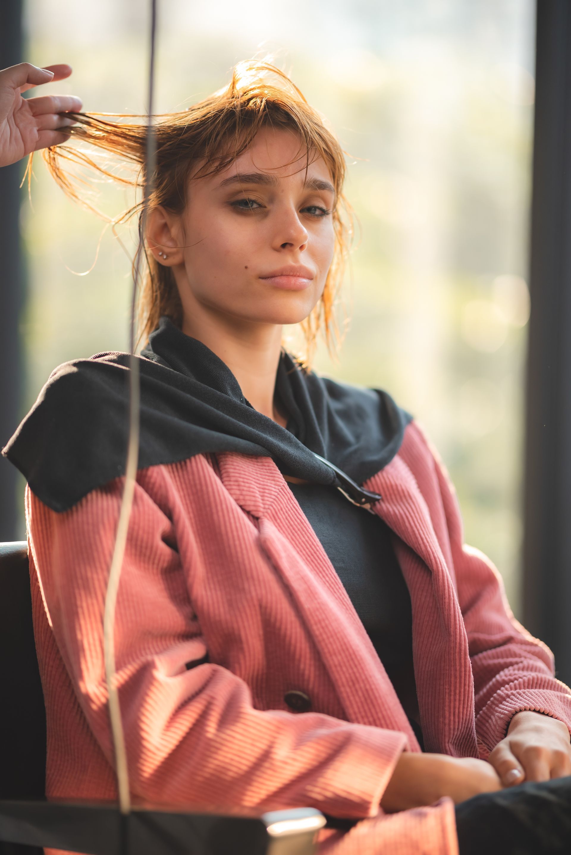 Woman in salon chair, hair being styled, wearing pink robe, sunlight, looking at camera.