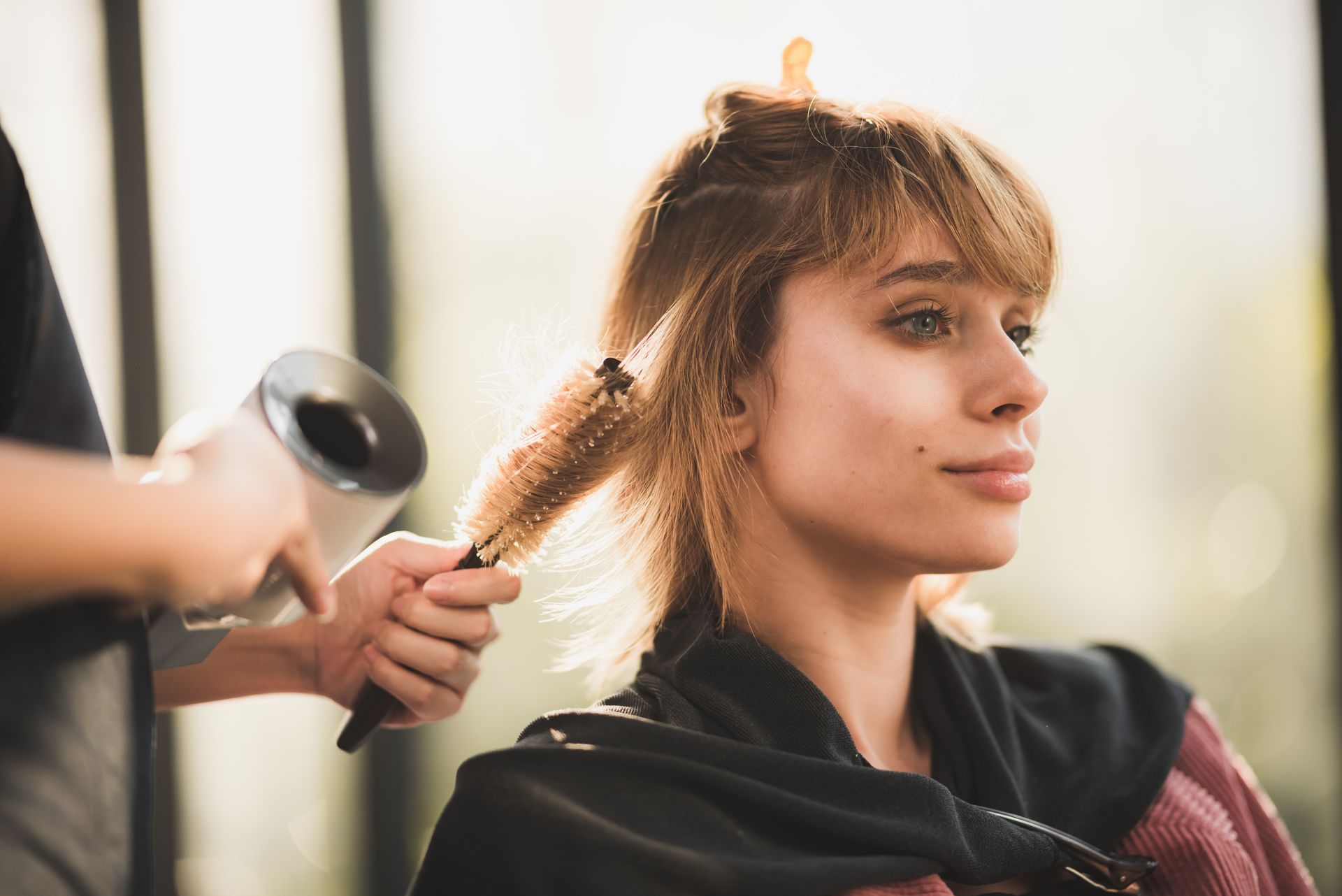 Woman getting her hair styled with a round brush and hairdryer at a salon.
